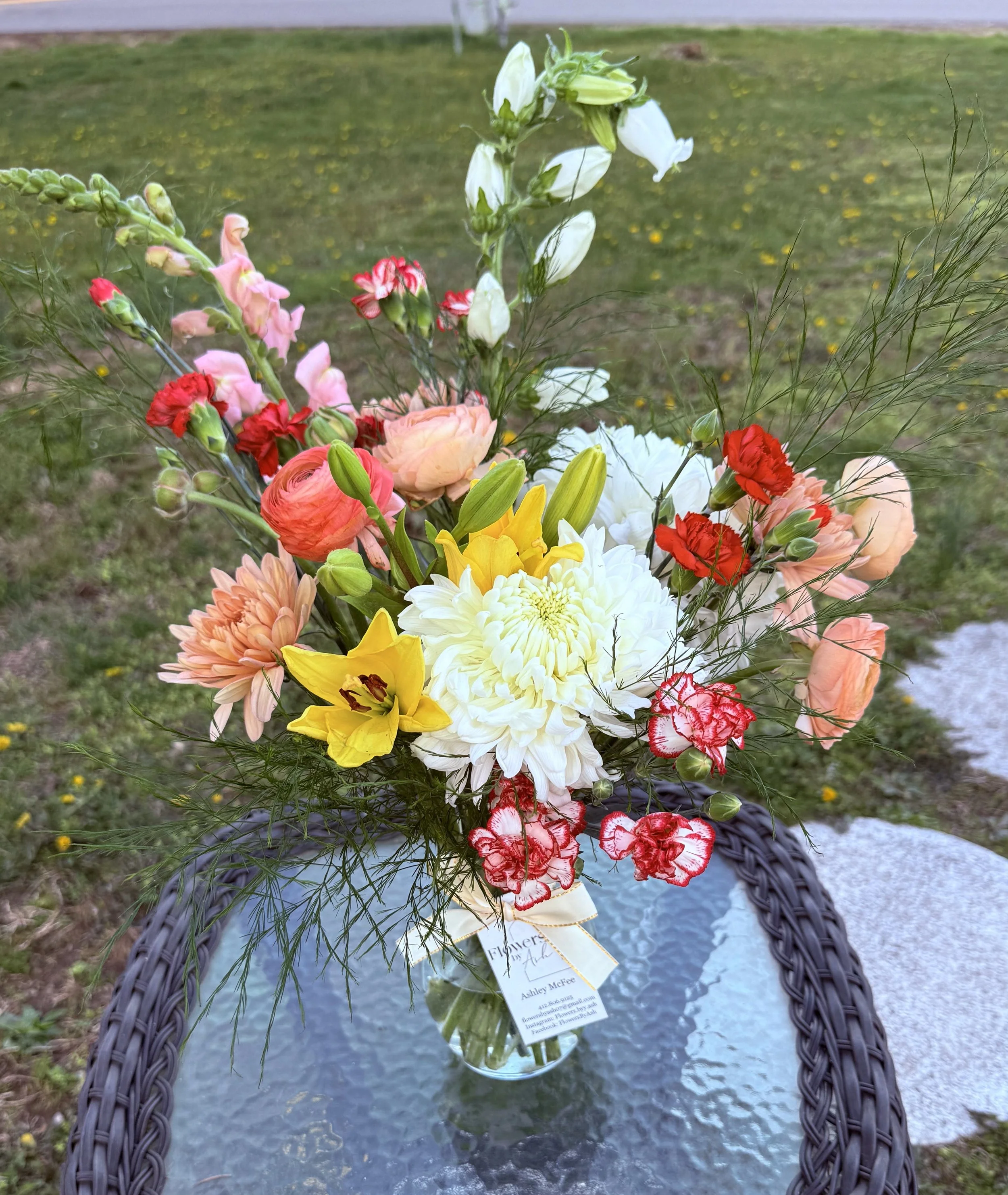 Colorful flower bouquet in a clear vase on a wicker table, featuring yellow daffodils, white and peach chrysanthemums, red carnations, and pink ranunculus, with greenery, against a grassy background.
