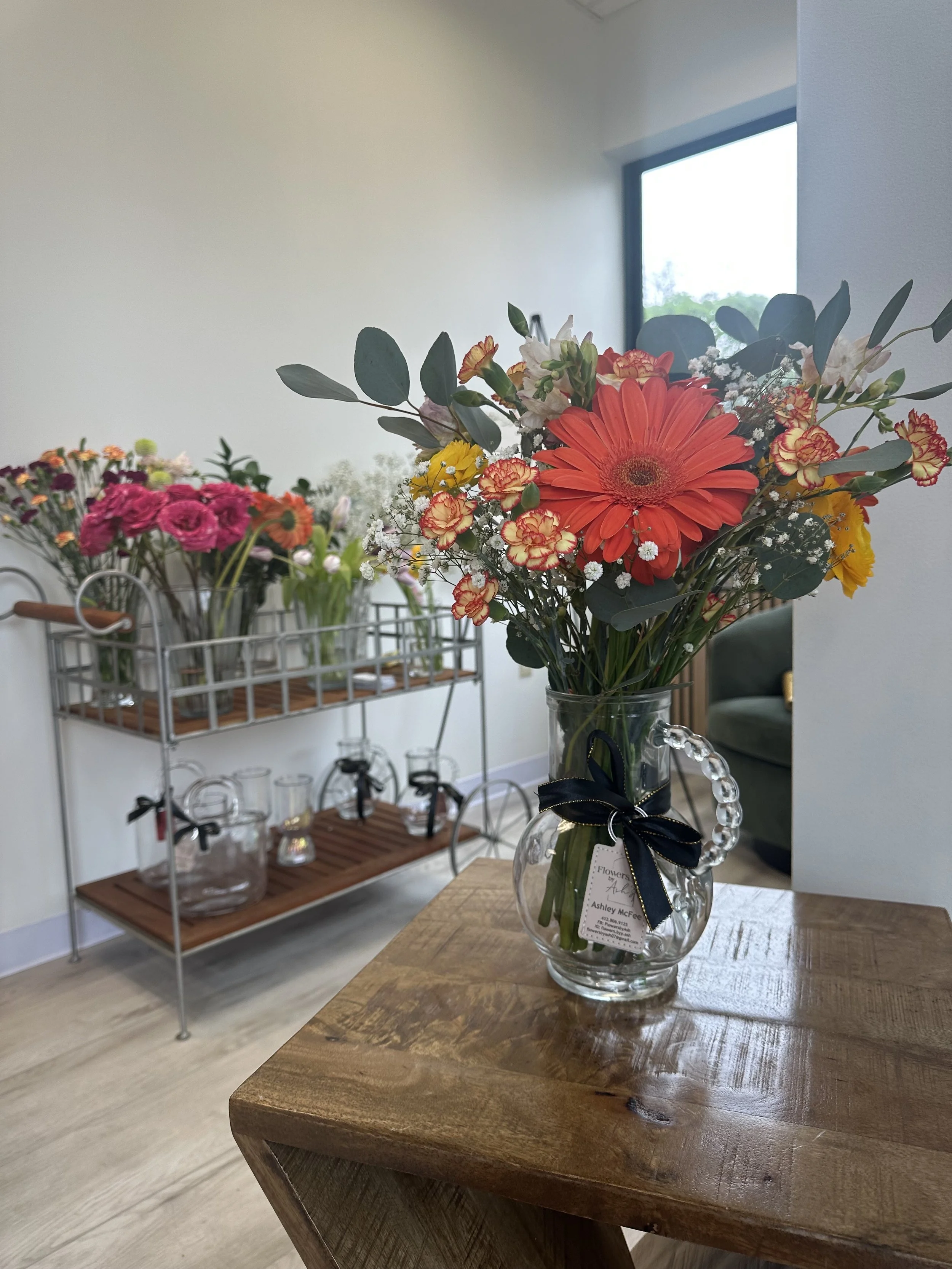 Bouquet of mixed flowers in a vase on a wooden table, with additional flower arrangements on a metal cart in the background.