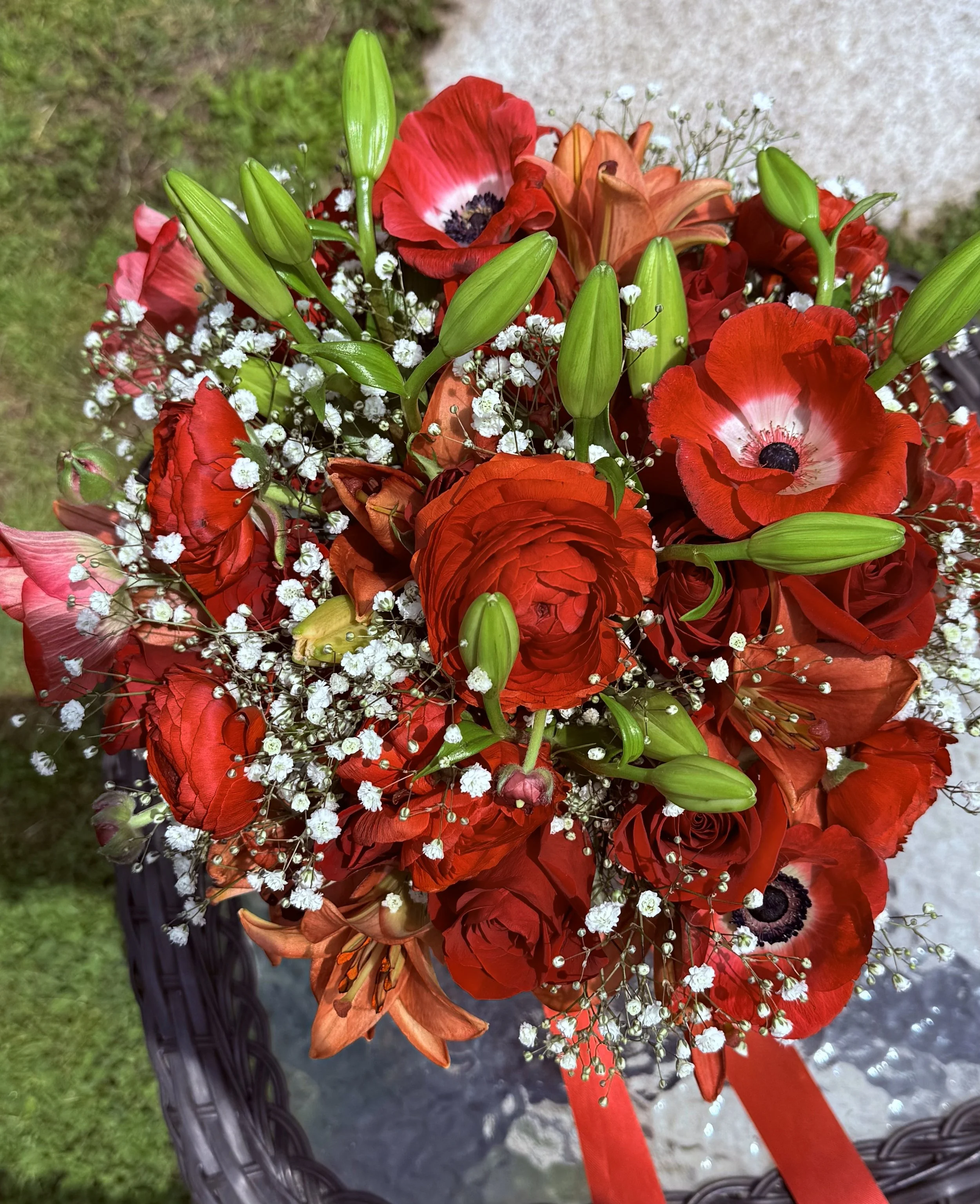 A bouquet of red roses, orange lilies, white baby’s breath, and green lily buds on a wicker table outdoors.