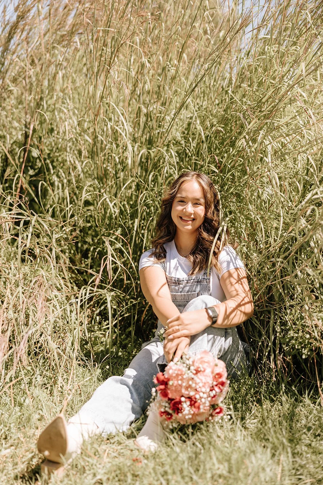 A young woman sitting among tall grass on a sunny day, holding a bouquet of pink and white flowers, smiling.