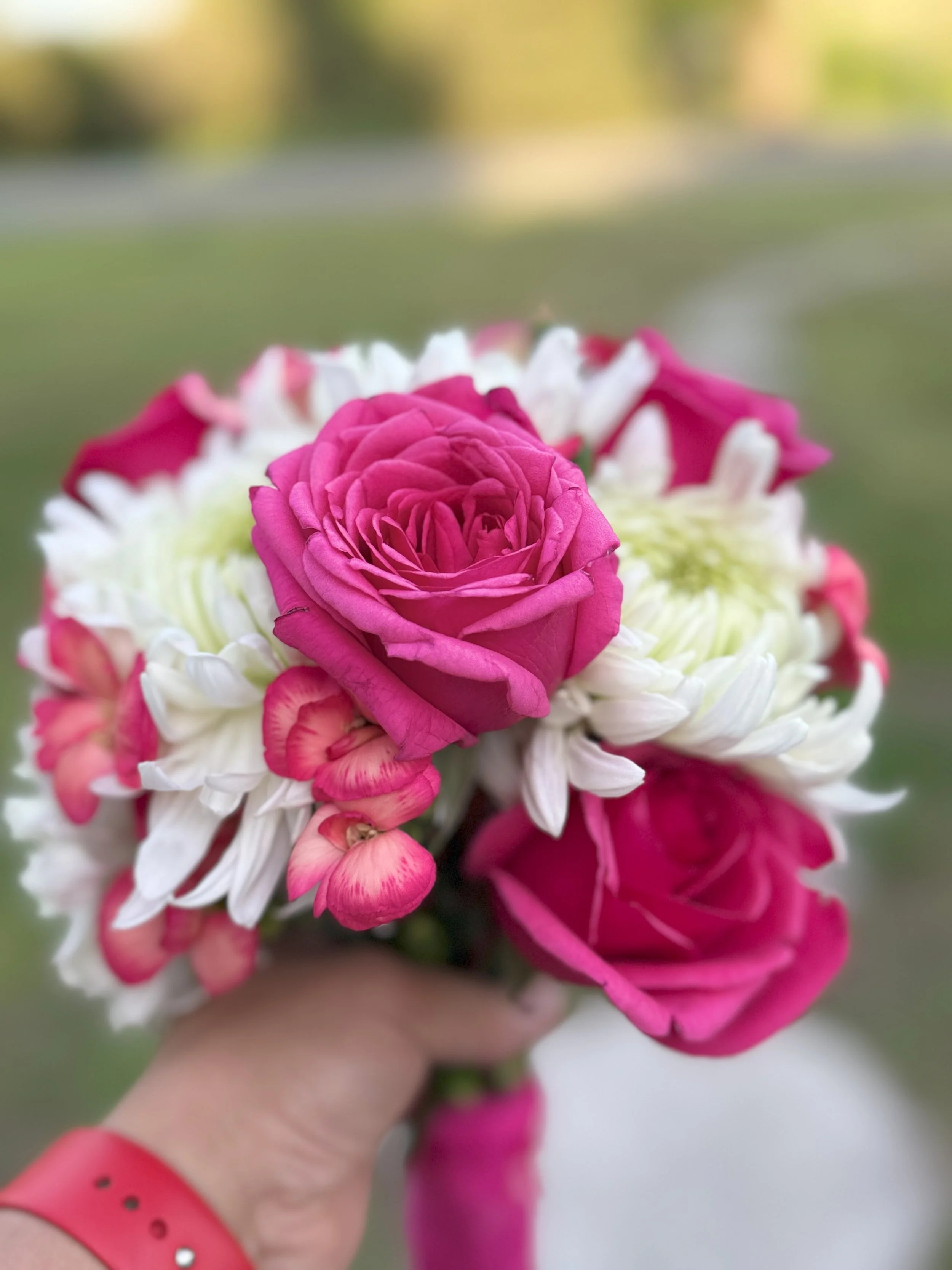 Close-up of a bouquet with pink roses, white and green chrysanthemums, and pink flowers, held by a hand wearing a red wristband, against a blurred outdoor background.