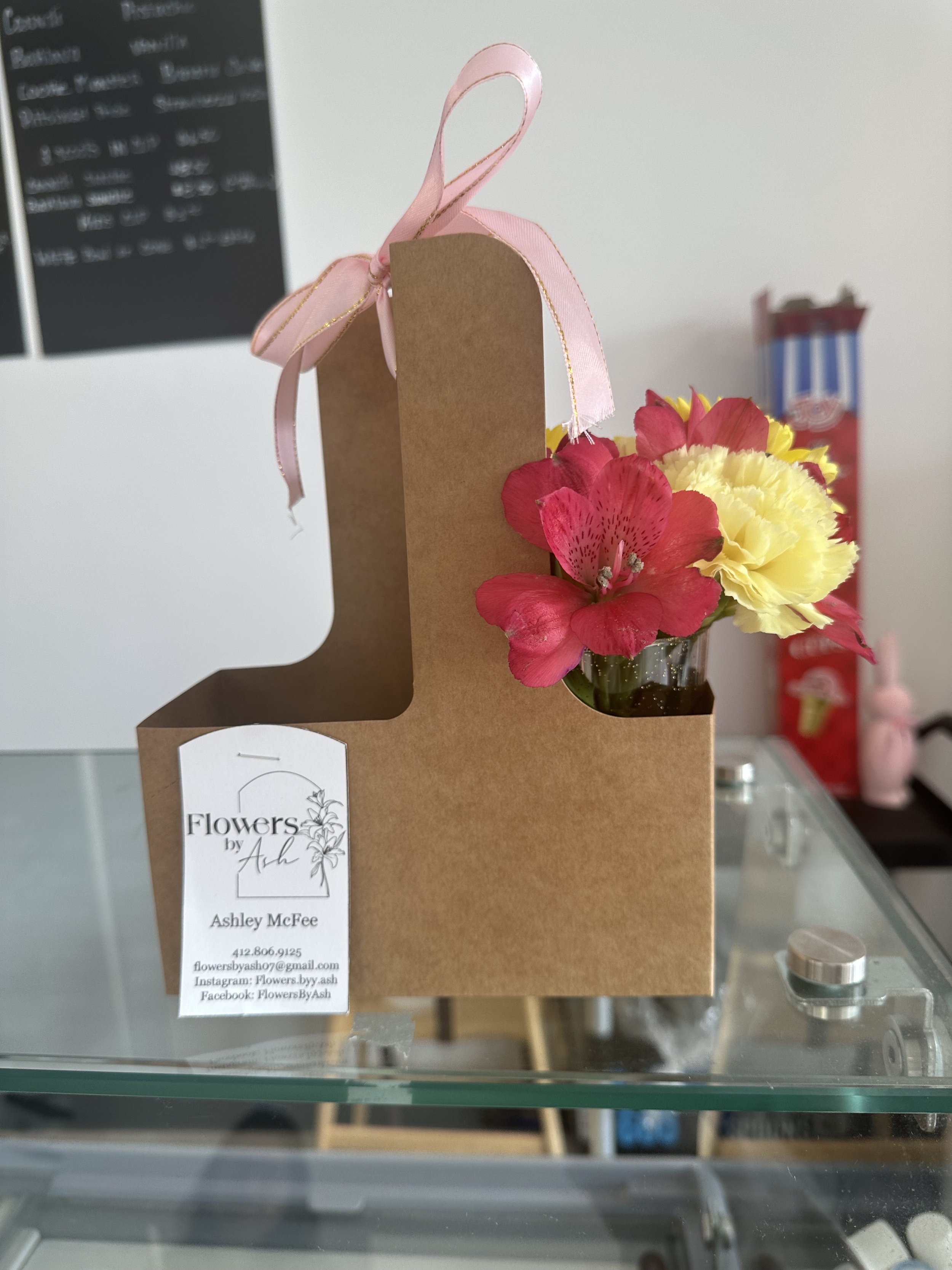 Flower arrangement in brown cardboard holder with pink ribbon, featuring pink and yellow flowers, and a business card from 'Flowers by Ash' on an indoor glass table.