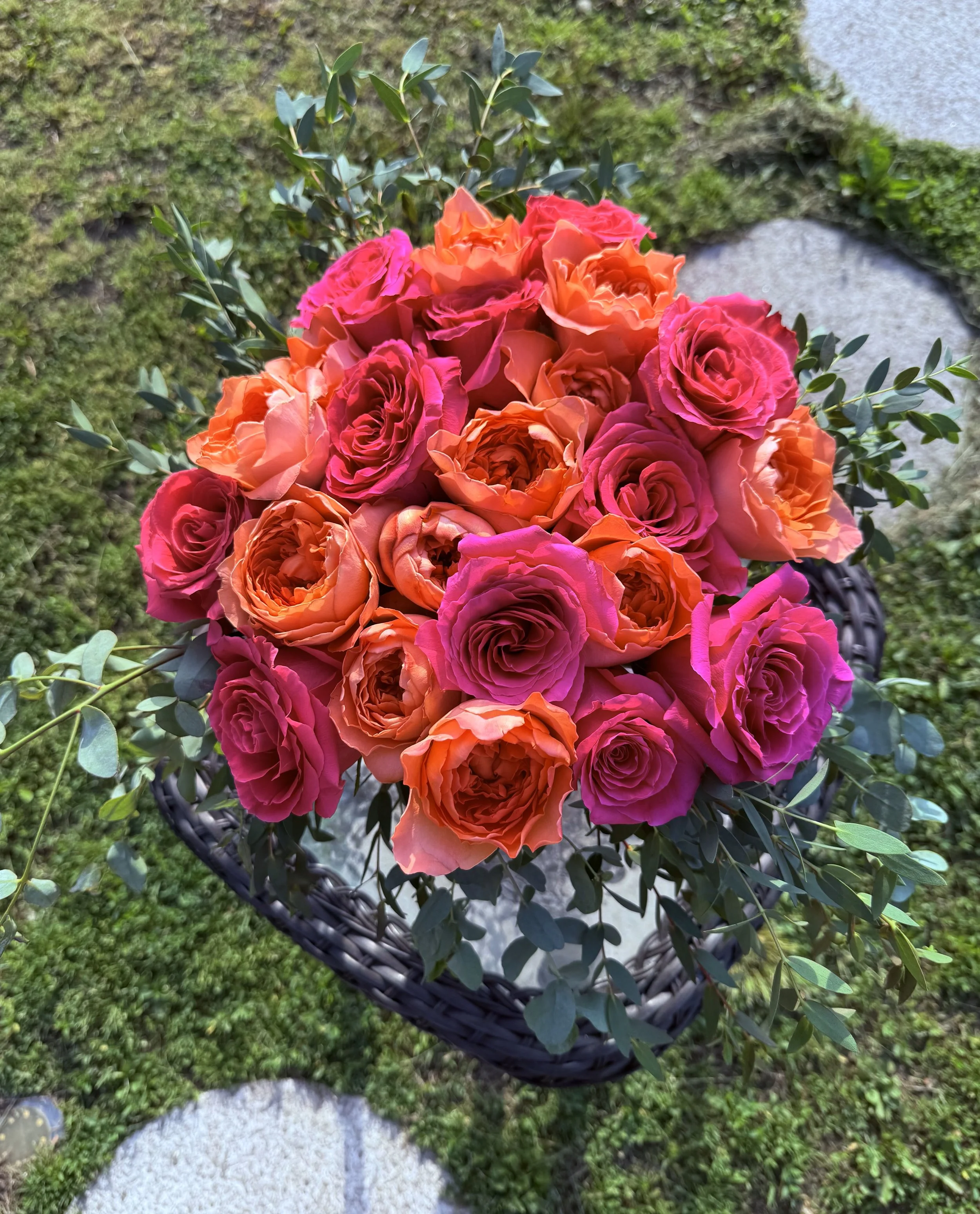 The image depicts a vibrant bouquet of roses in shades of pink and coral, arranged with eucalyptus leaves in a black wicker basket. The arrangement is placed on a moss-covered surface, adding a touch of natural elegance to the overall scene.