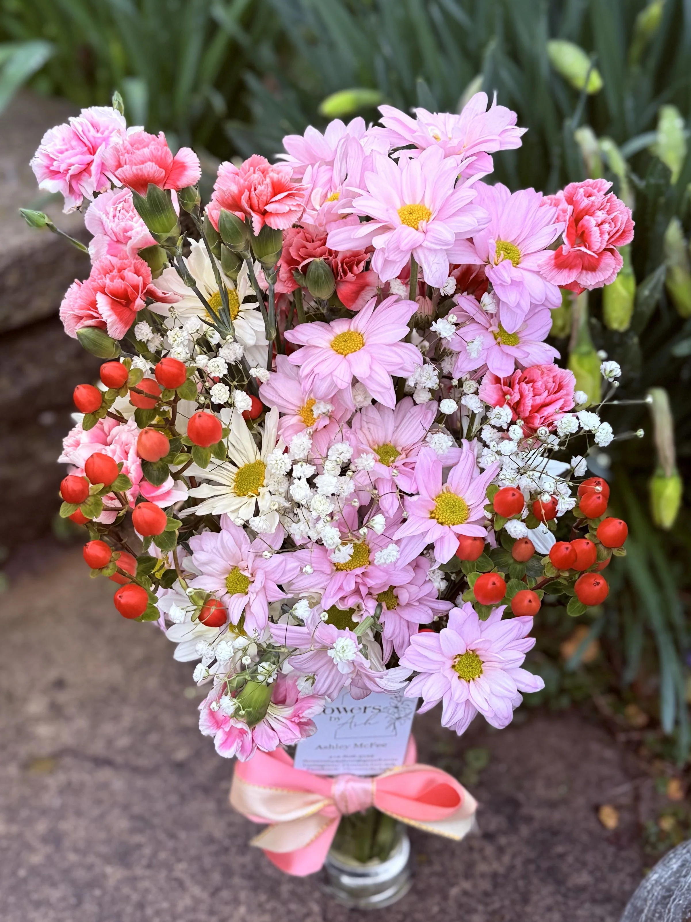 A bouquet featuring pink daisies, pink carnations, baby's breath, and red berry accents in a glass vase, tied with a pink ribbon, set outdoors.