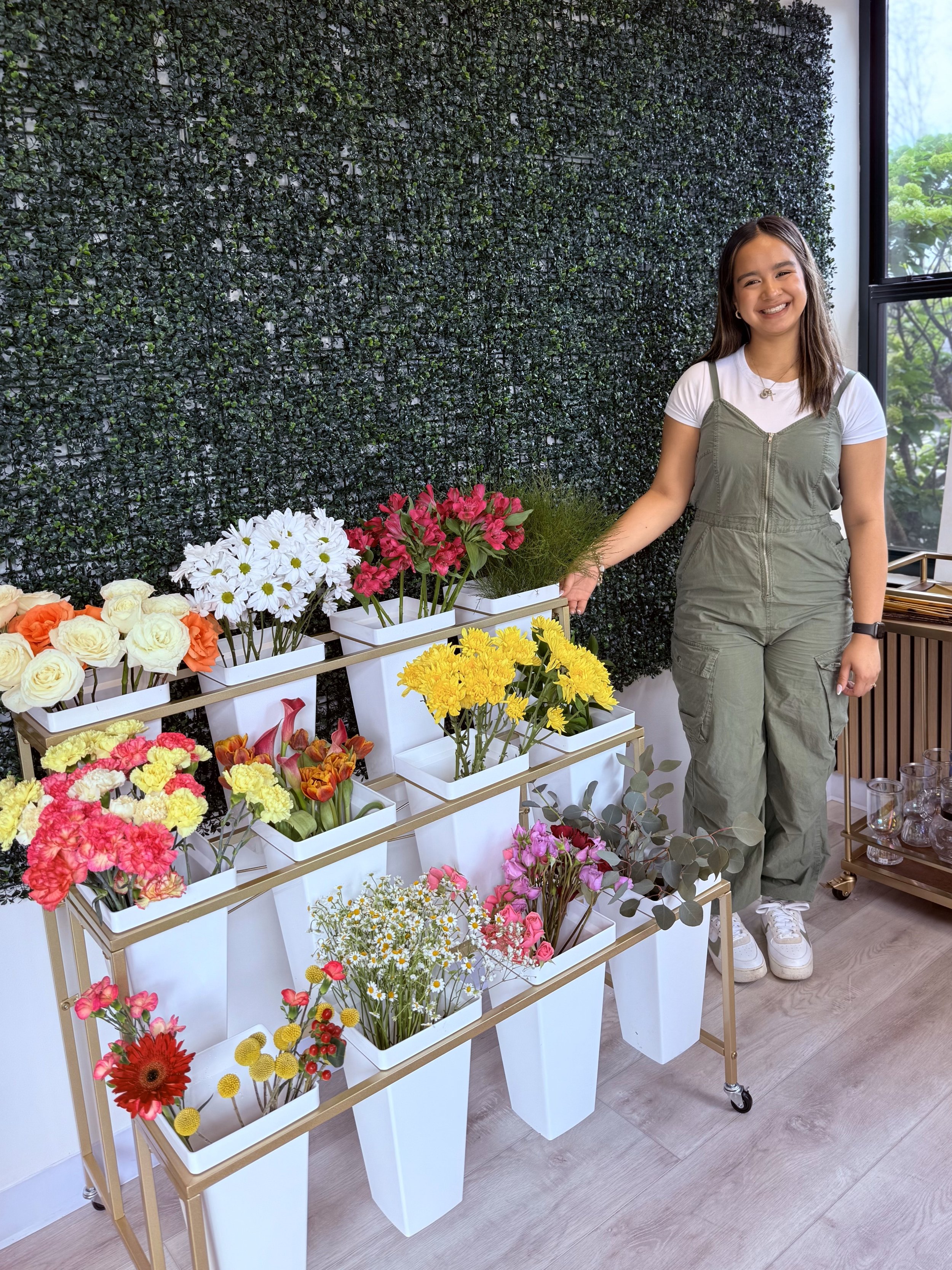 Young woman standing next to a display of colorful flower arrangements in white vases with a green wall backdrop and a large window.
