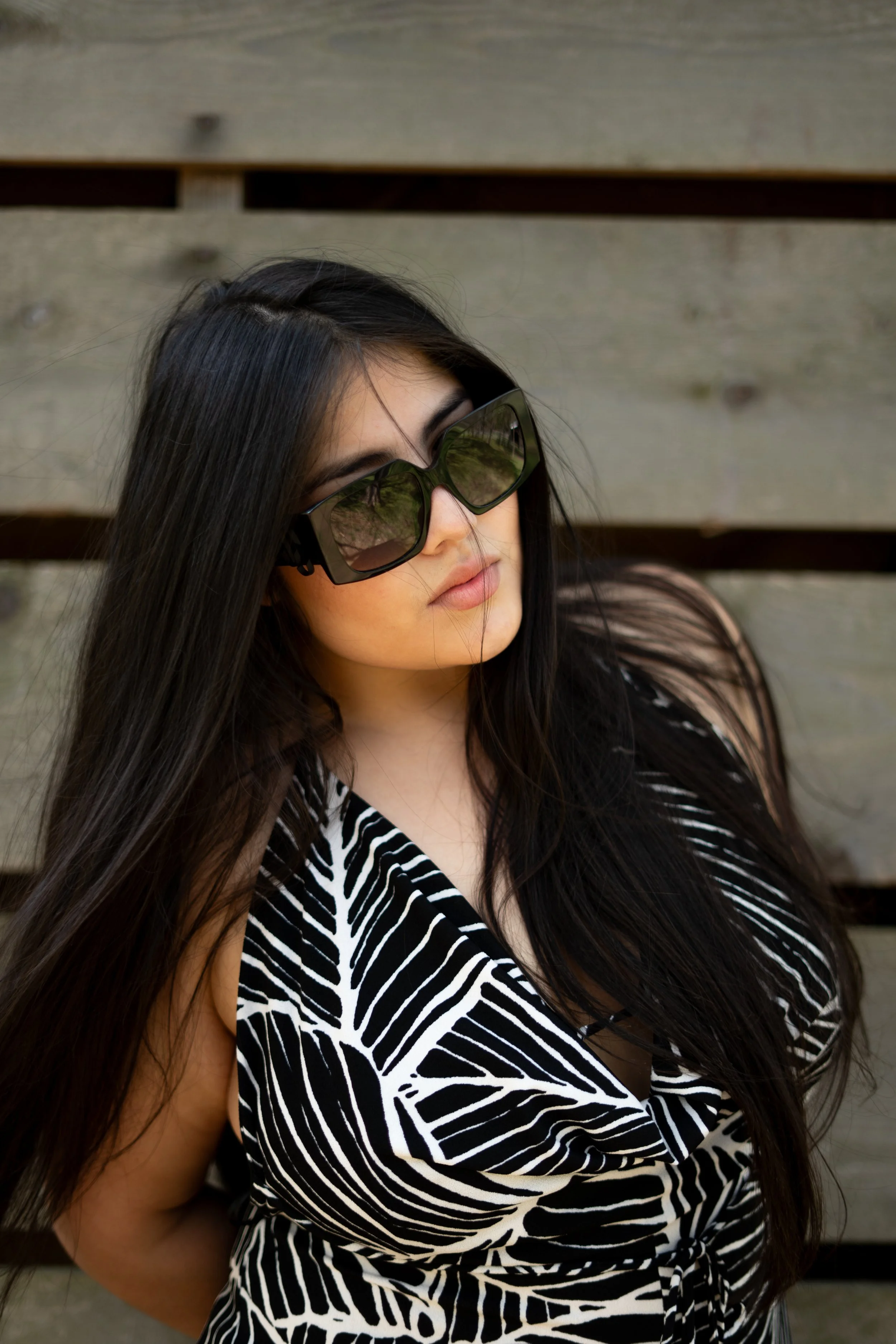 A woman with long dark hair wearing black sunglasses and a black and white zebra-patterned top, standing in front of a wooden background.
