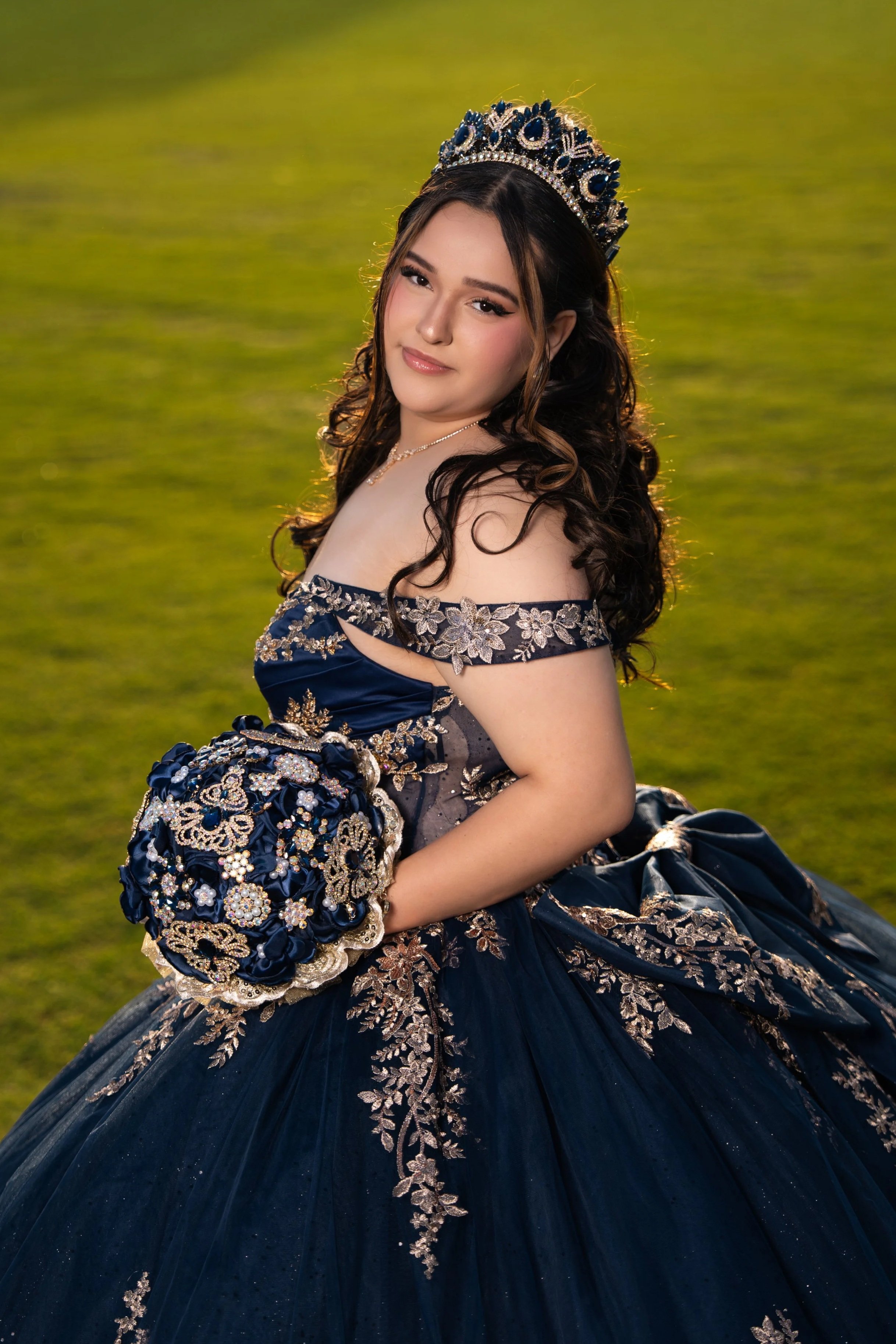 Young woman in a dark blue, embroidered gown with a matching tiara, holding a bouquet of flowers, outdoors on a grassy field.