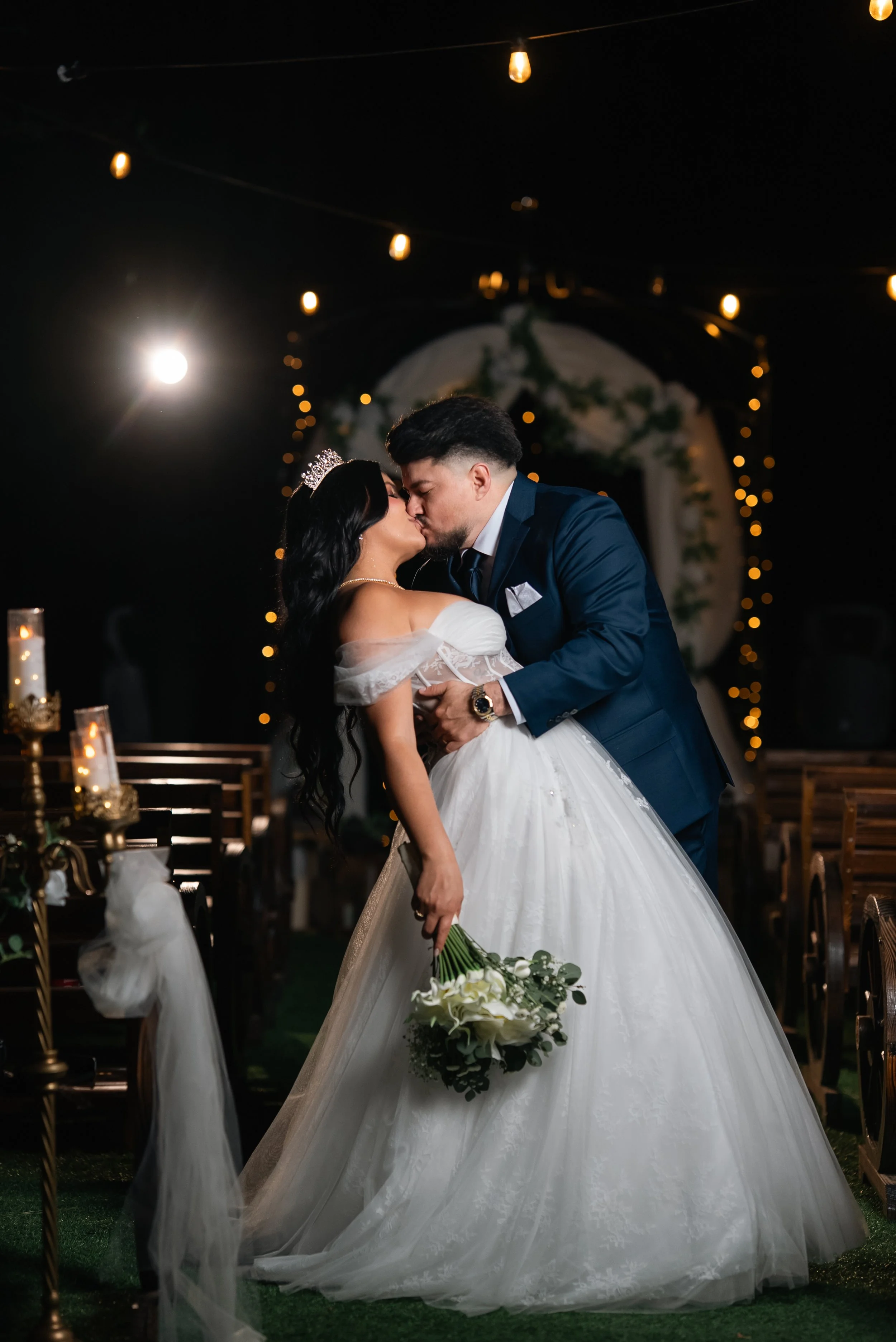 A bride and groom sharing a kiss during their wedding ceremony, with wedding decorations and string lights in the background.