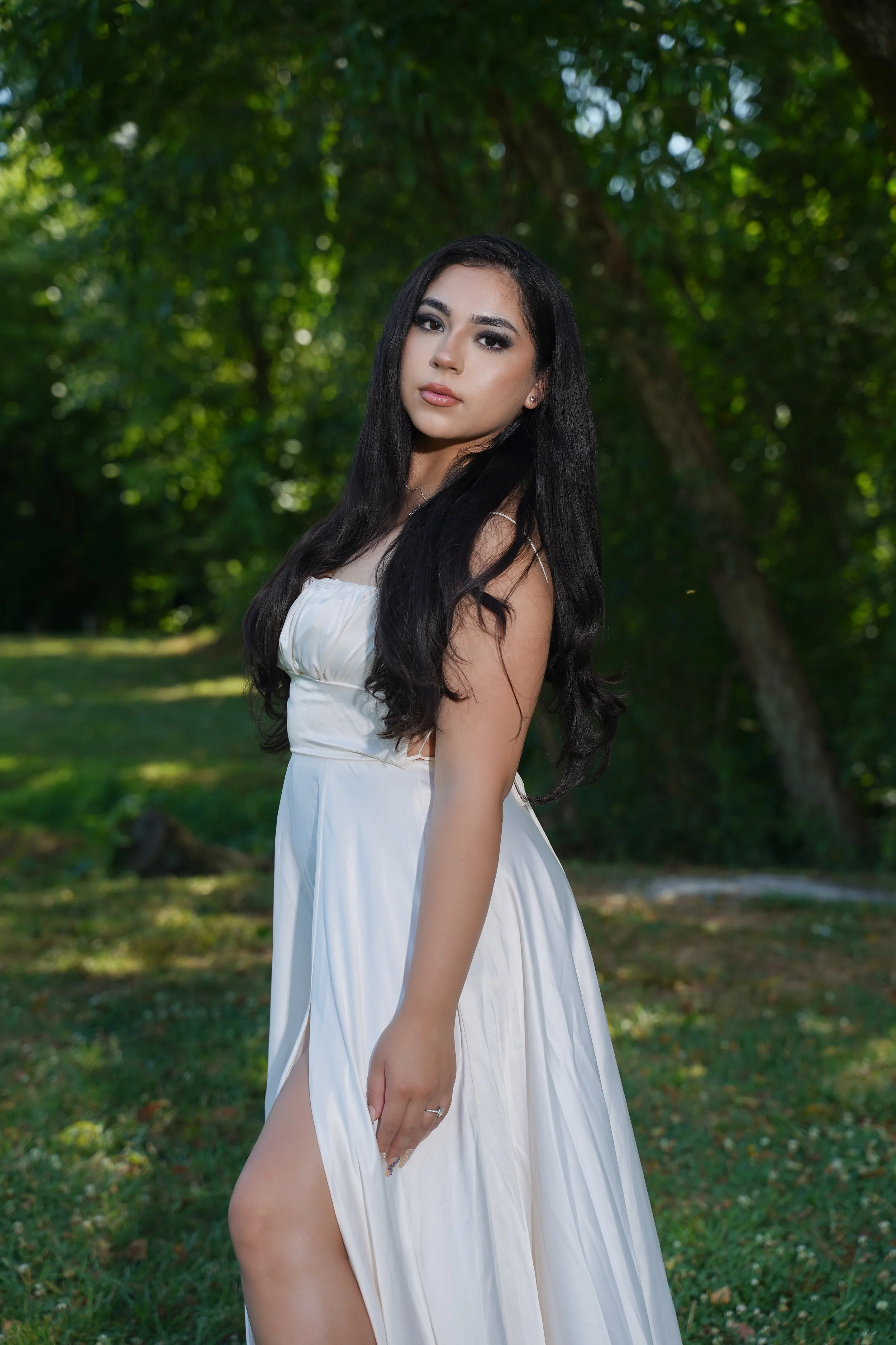 A woman with long black hair wearing a white dress standing outdoors in a lush green park with trees in the background.
