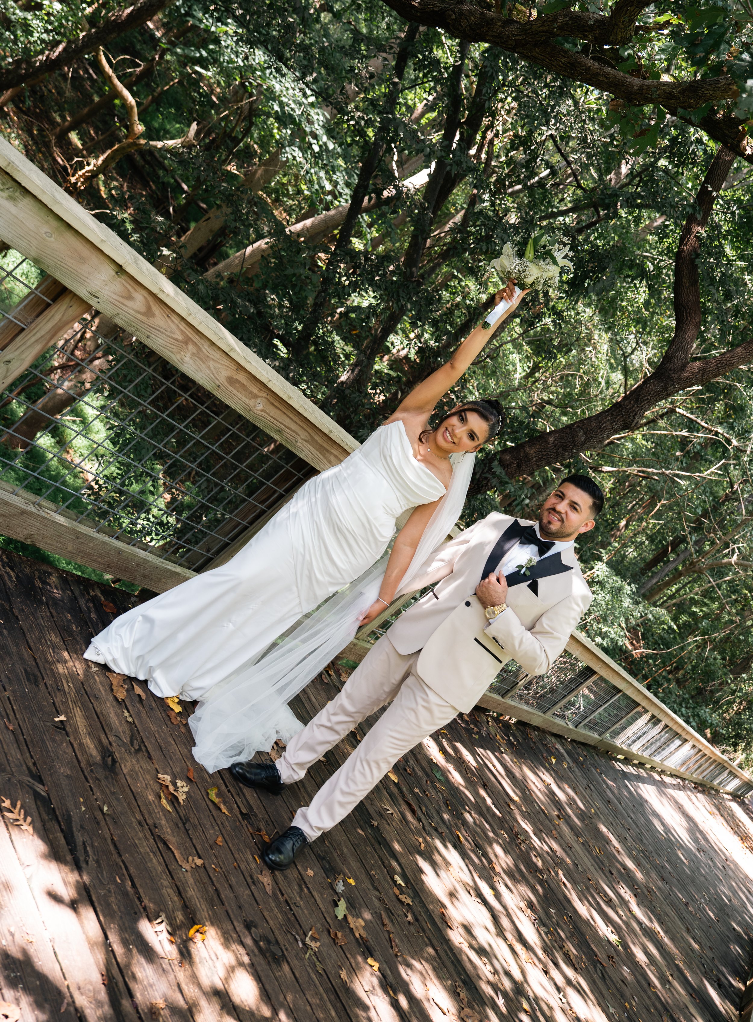 A bride and groom standing on a wooden pathway in a wooded area, with the bride holding a bouquet of white flowers and the groom wearing a cream tuxedo with a black bow tie. She is smiling, and he is looking slightly to the side.