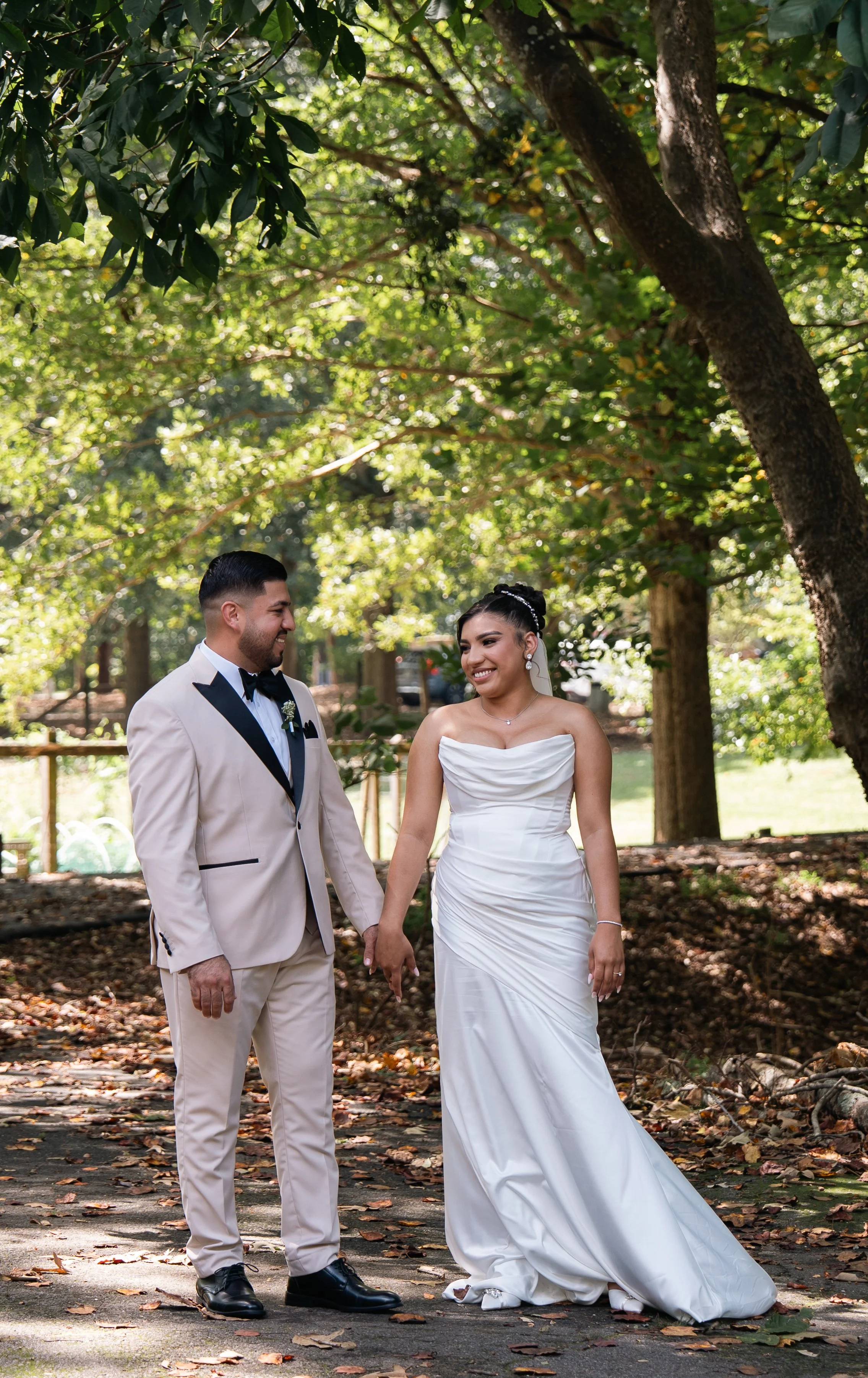 A bride and groom stand hand-in-hand outdoors in a wooded area during daytime, smiling at each other, with sunlight filtering through the trees.