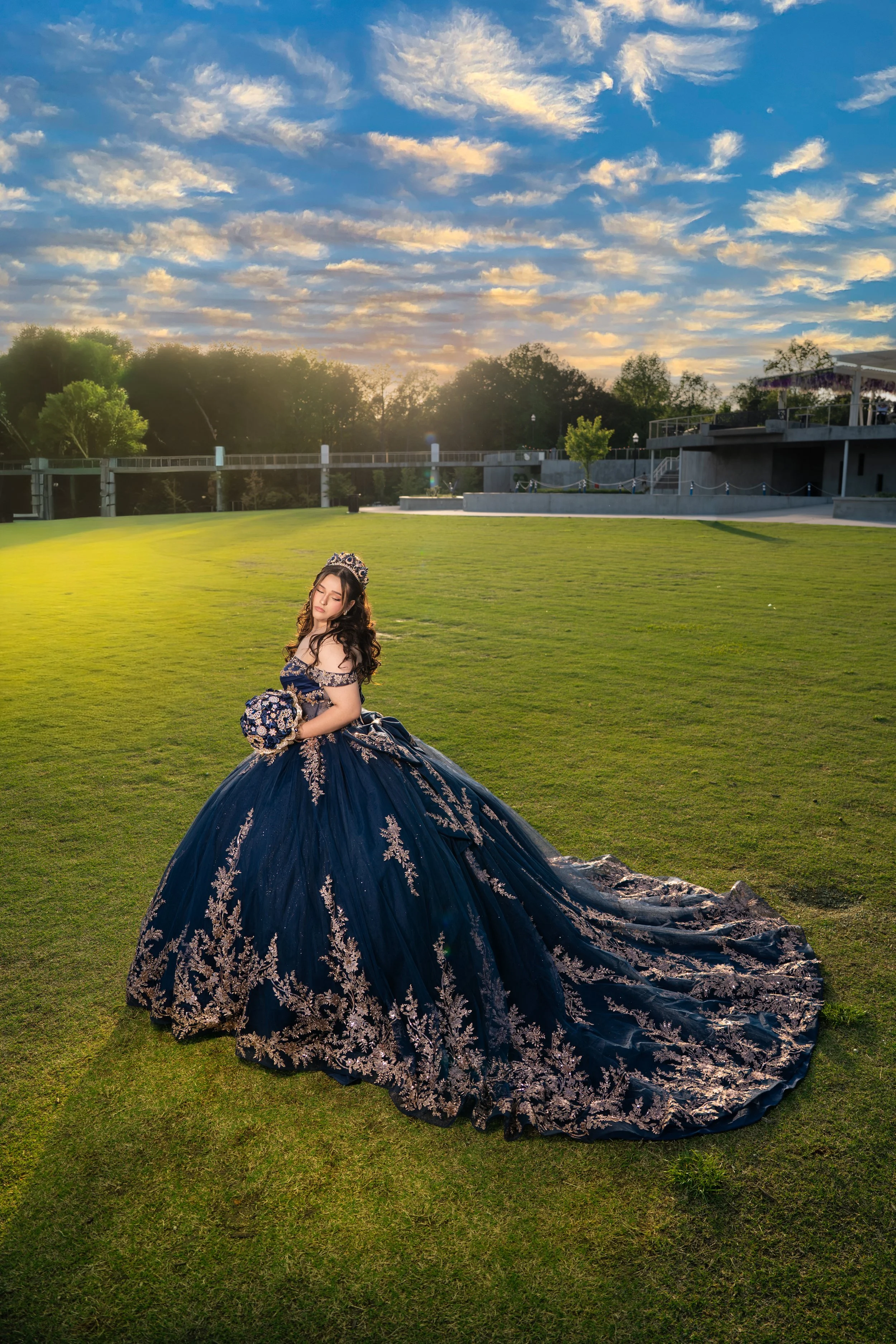 A young woman wearing a dark blue ball gown with intricate gold embroidery, sitting on the grass in an outdoor park during sunset. The sky has scattered clouds with golden and blue hues, and the background features trees, a building, and a deck.