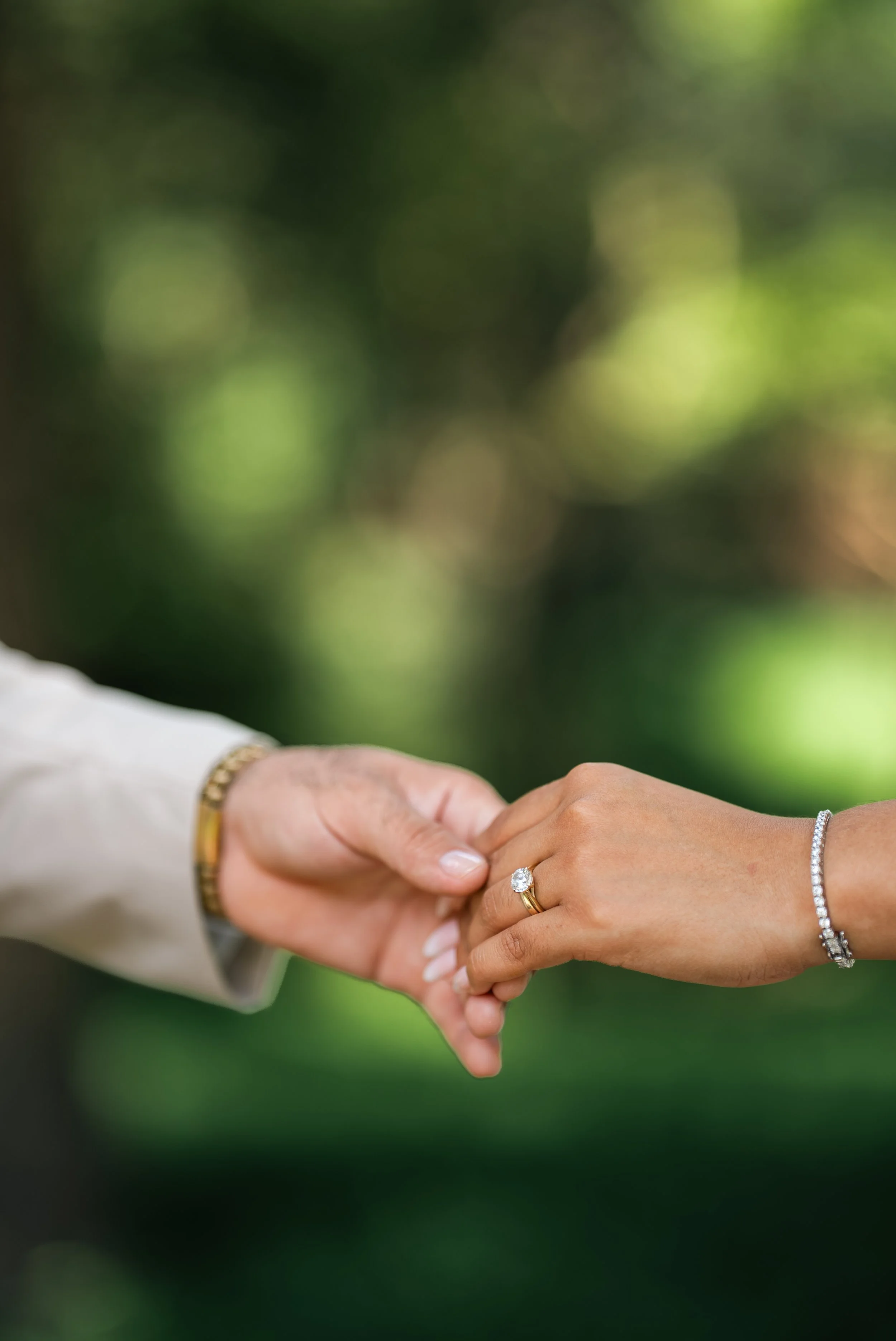 A close-up of a couple holding hands, with the woman wearing a ring and bracelet, and the man wearing a watch, against a blurred green outdoor background.
