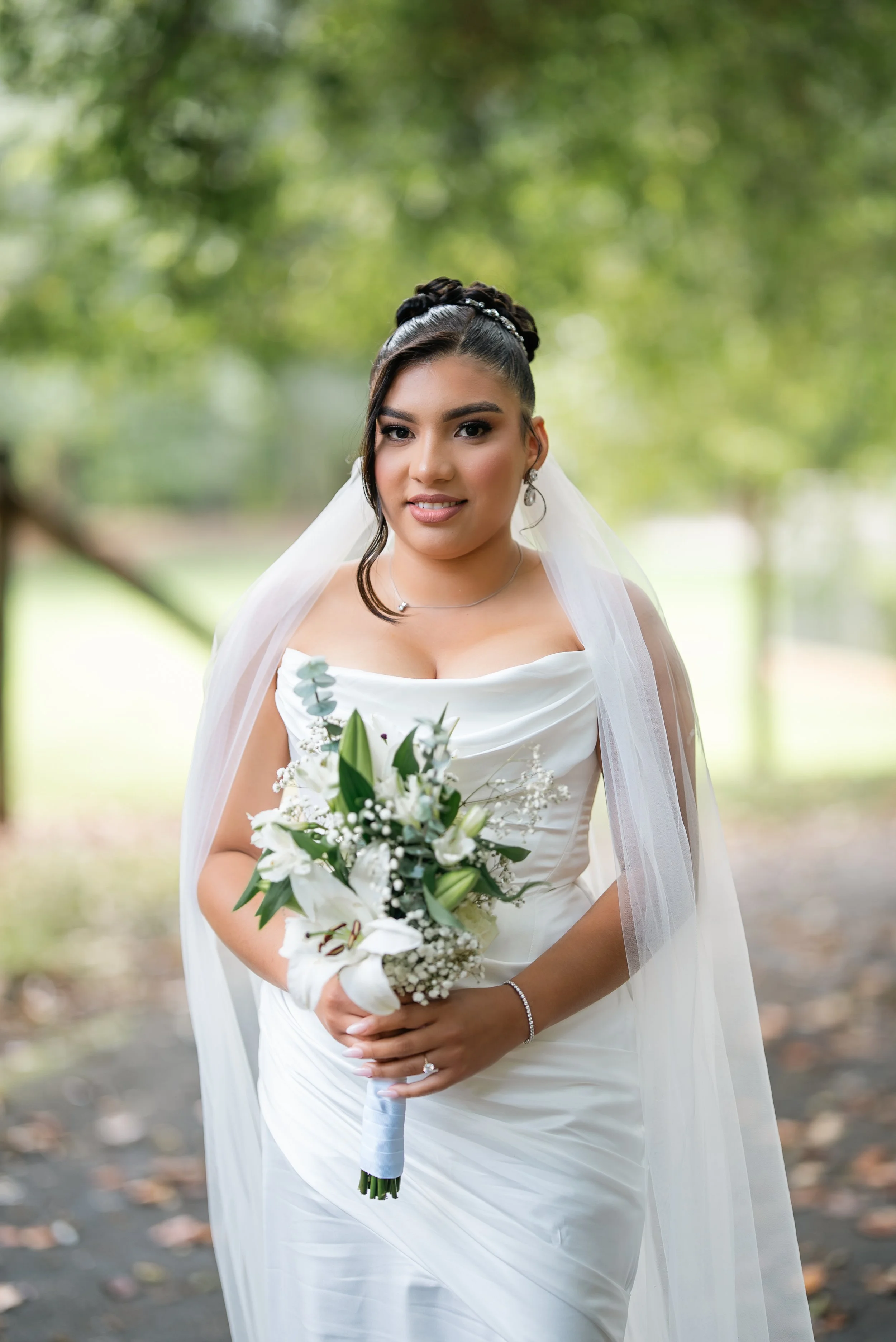 A bride in a white wedding dress holding a bouquet of white lilies and greenery, standing outdoors in a wooded area, smiling at the camera.