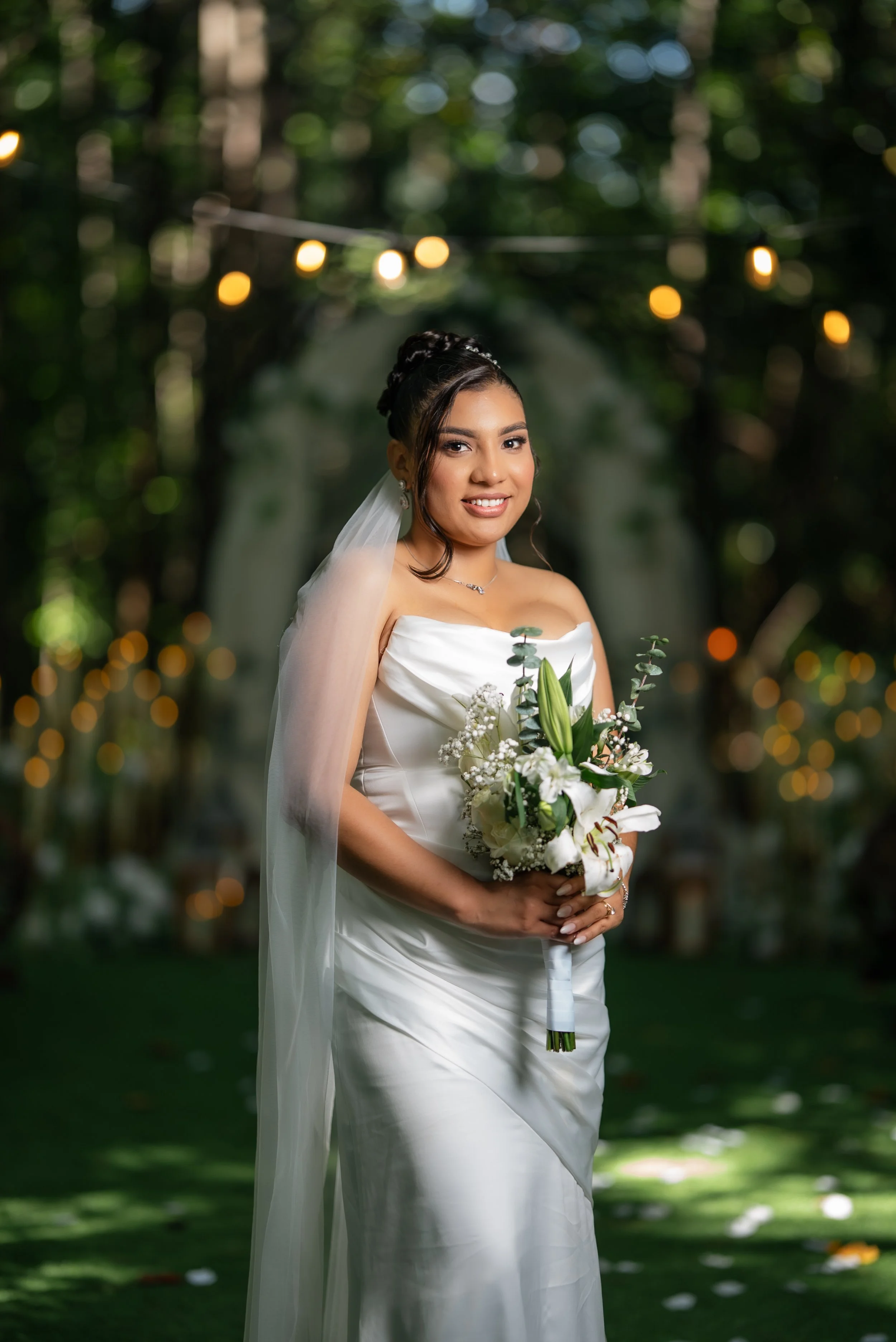A bride in a white strapless wedding gown holding a bouquet of white flowers, standing in a wooded outdoor setting with string lights overhead and a blurred green background.