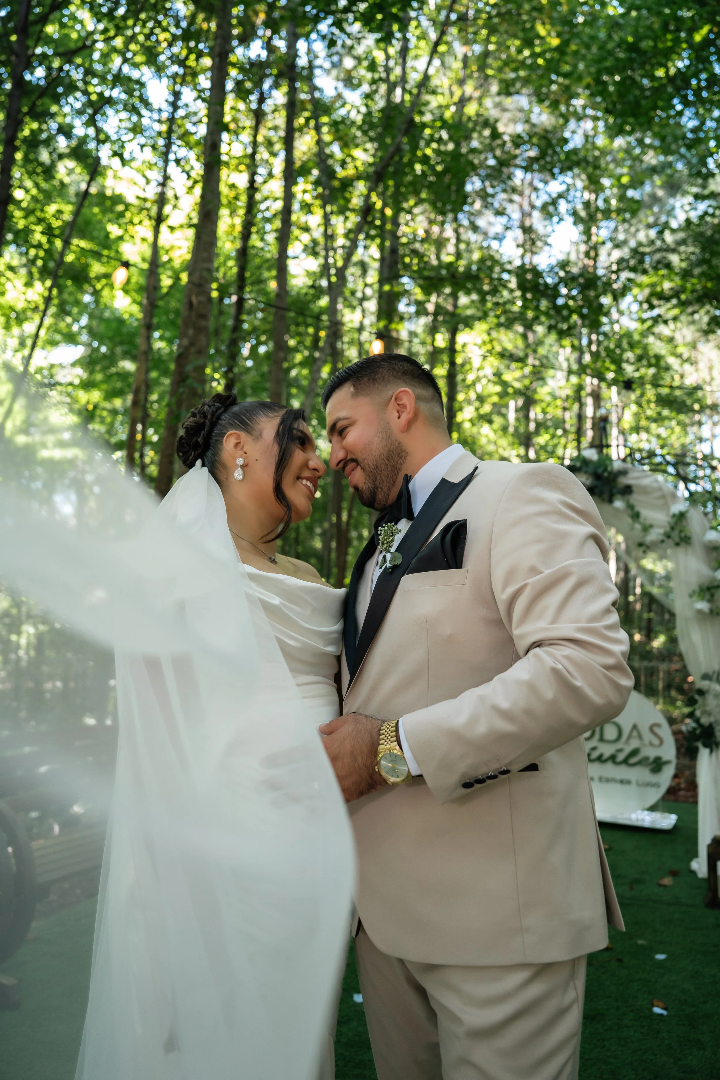 A bride and groom are smiling and touching foreheads during a wedding ceremony outdoors among tall trees with green leaves, sunlight filtering through.