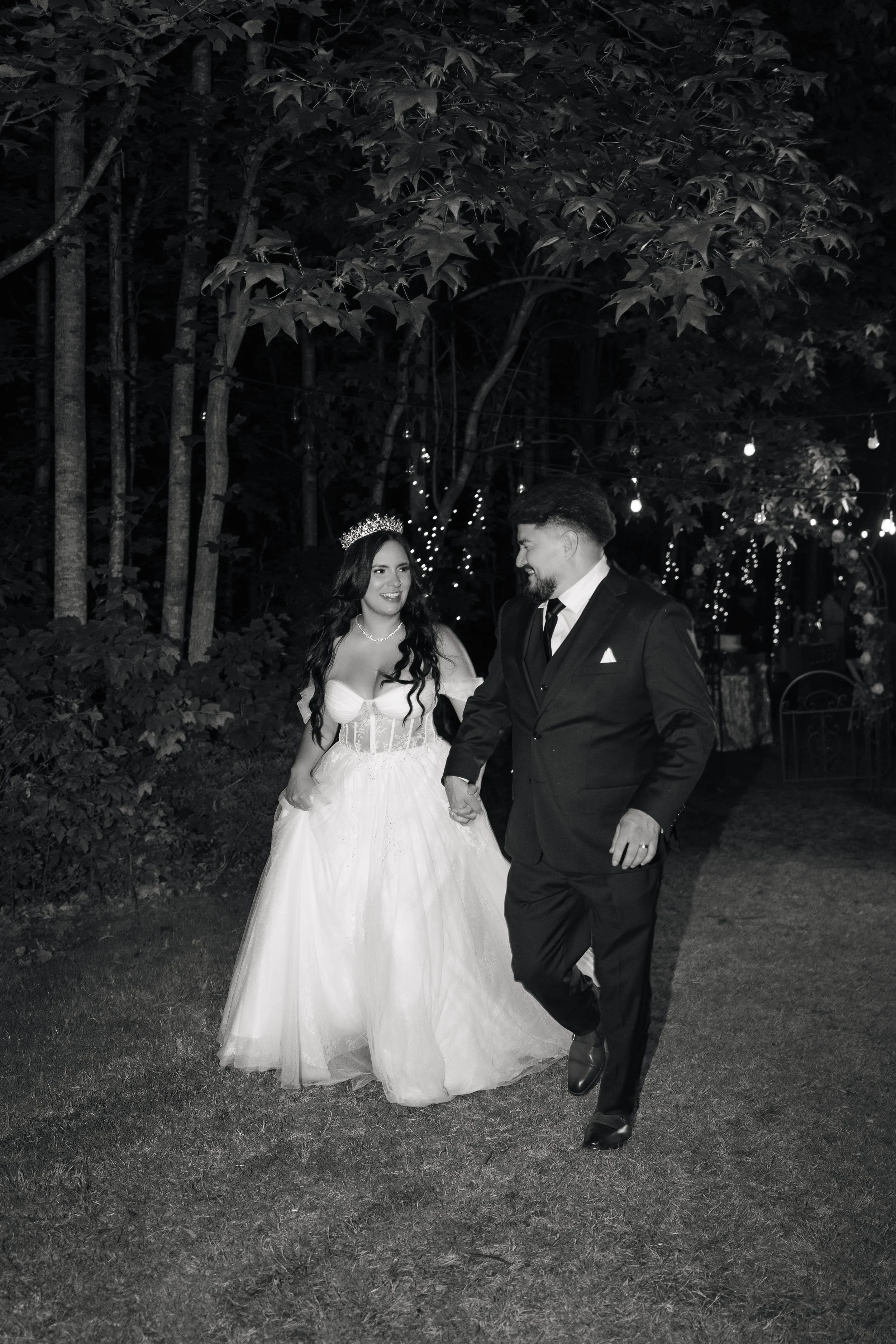 A bride and groom holding hands and smiling at each other during their wedding reception outdoors at night, with string lights hanging above and trees in the background.