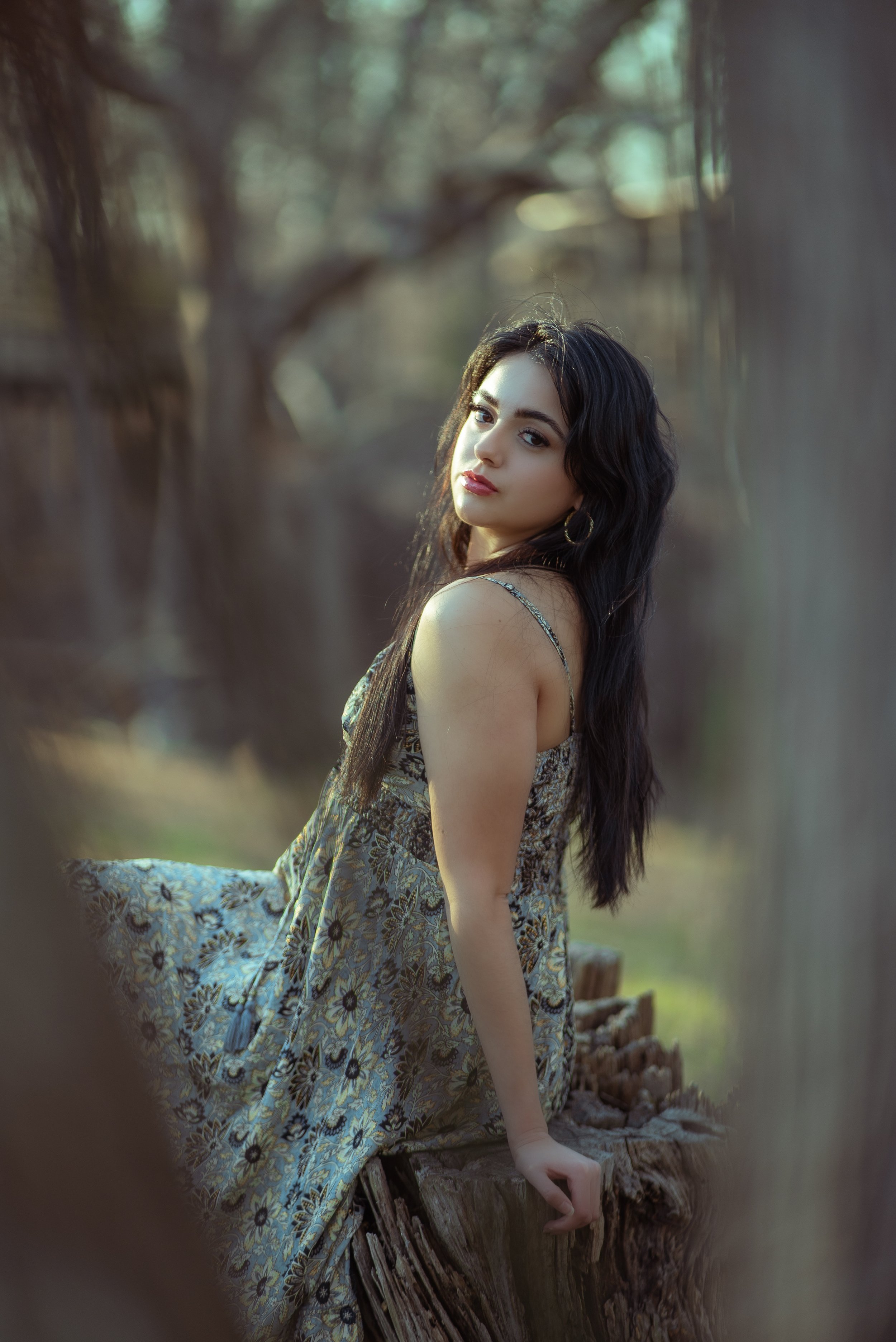 A young woman with long dark hair and hoop earrings sits on a weathered tree stump outdoors, wearing a floral dress. She looks over her shoulder at the camera with a neutral expression, surrounded by a blurred natural background with trees and soft sunlight.