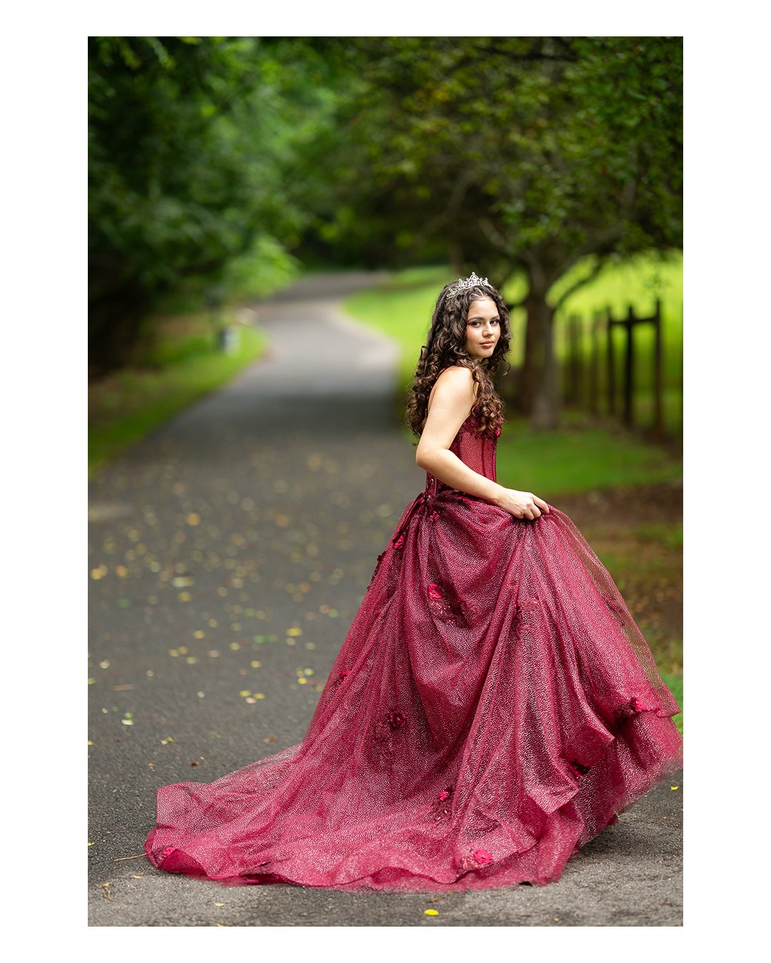 Young woman in a red gown and tiara standing on a road surrounded by trees.