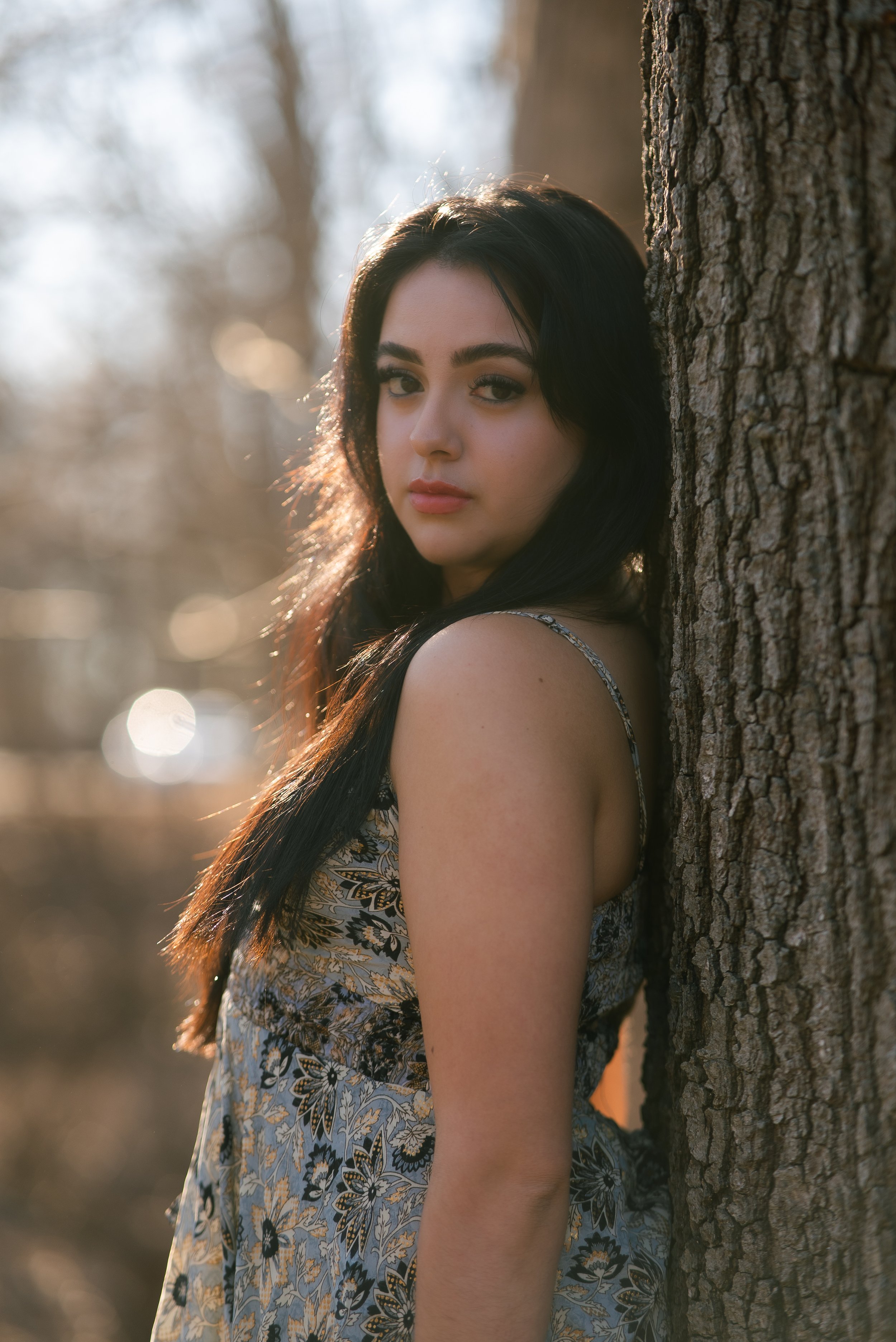 Young woman with long dark hair leaning against a tree in a forest during sunset.