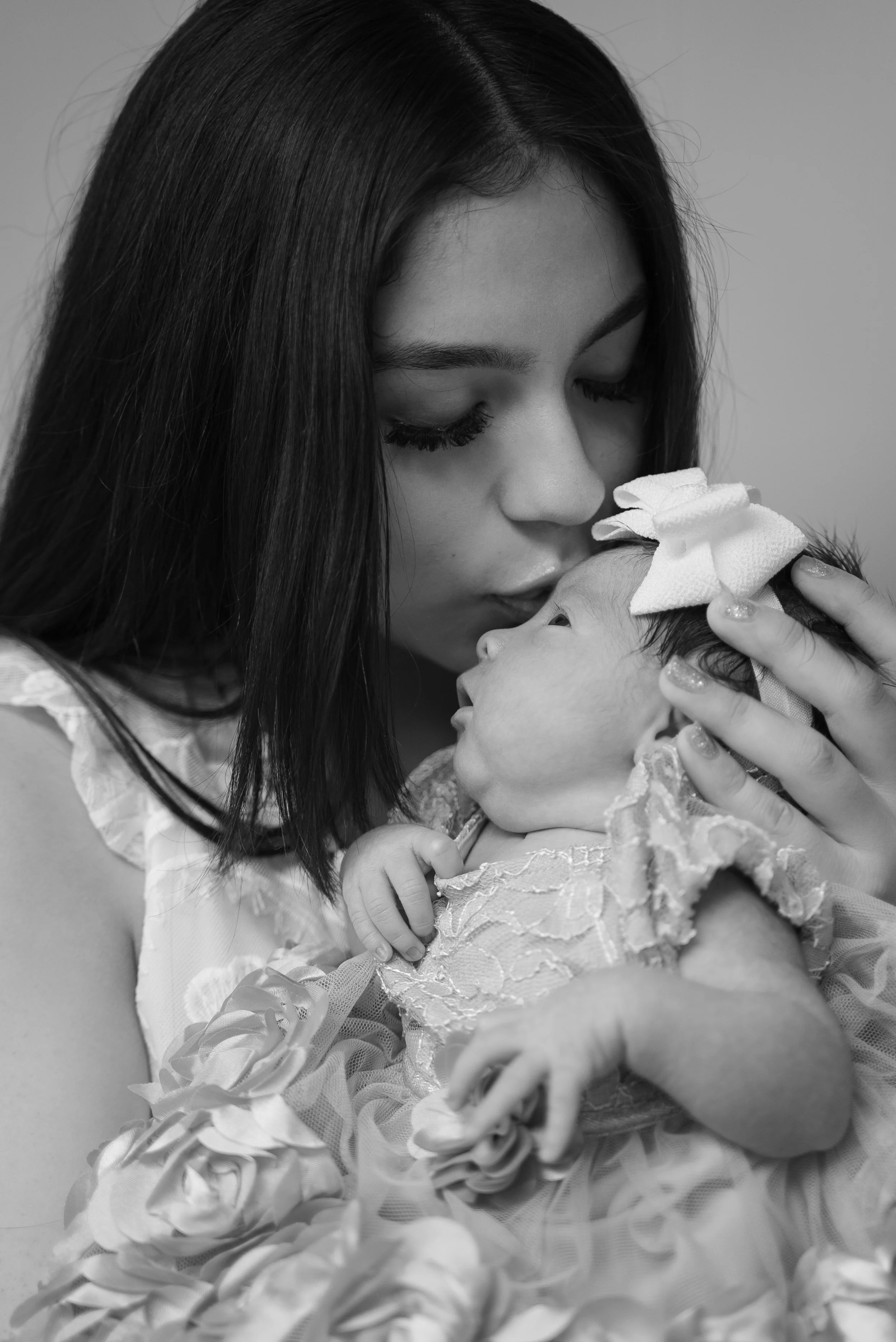 A woman holding and kissing a baby girl, both in floral dresses, in black and white.
