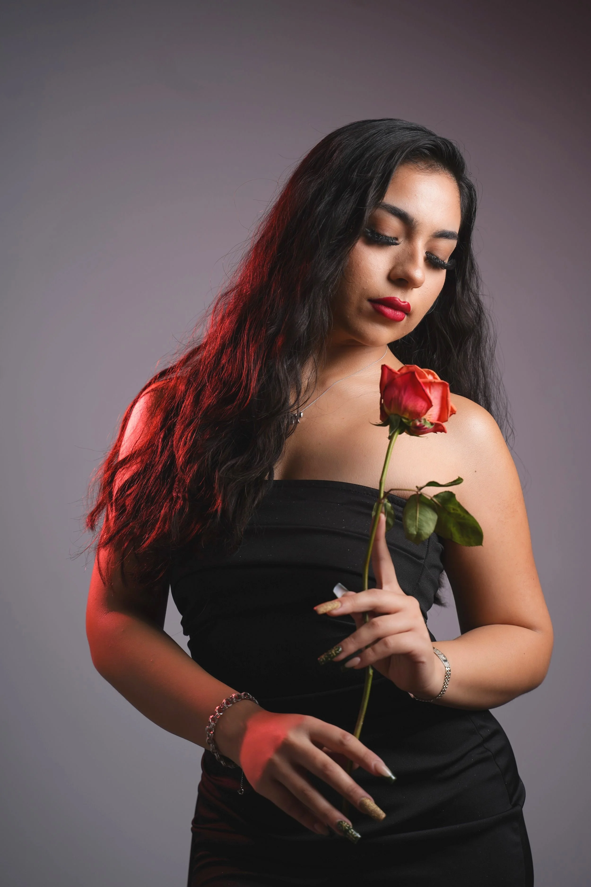 A woman with long dark wavy hair and red lipstick holding a red rose against a plain background.