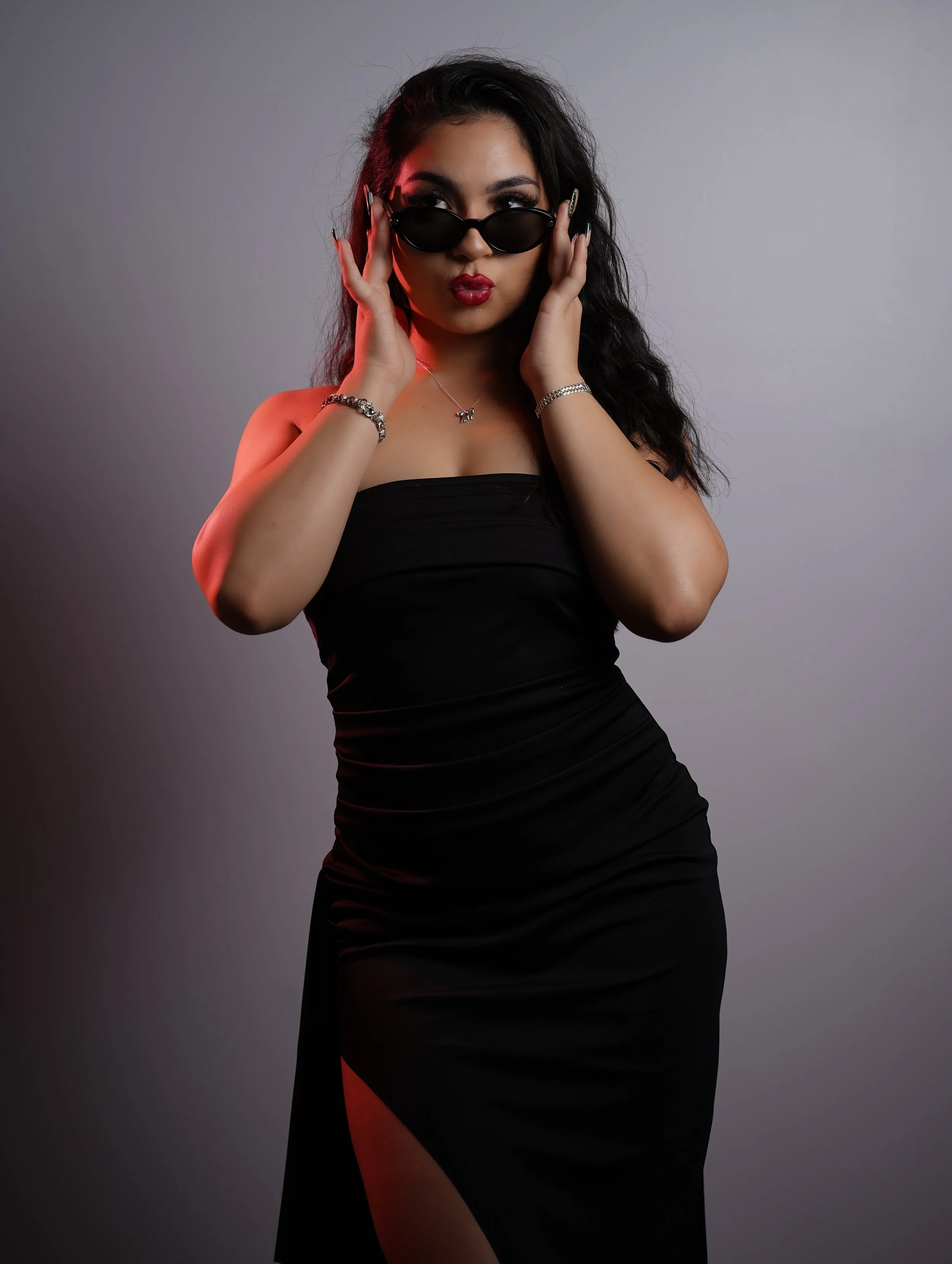 A woman with long dark hair in a black strapless dress and sunglasses, posing with her hands near her face against a plain background.