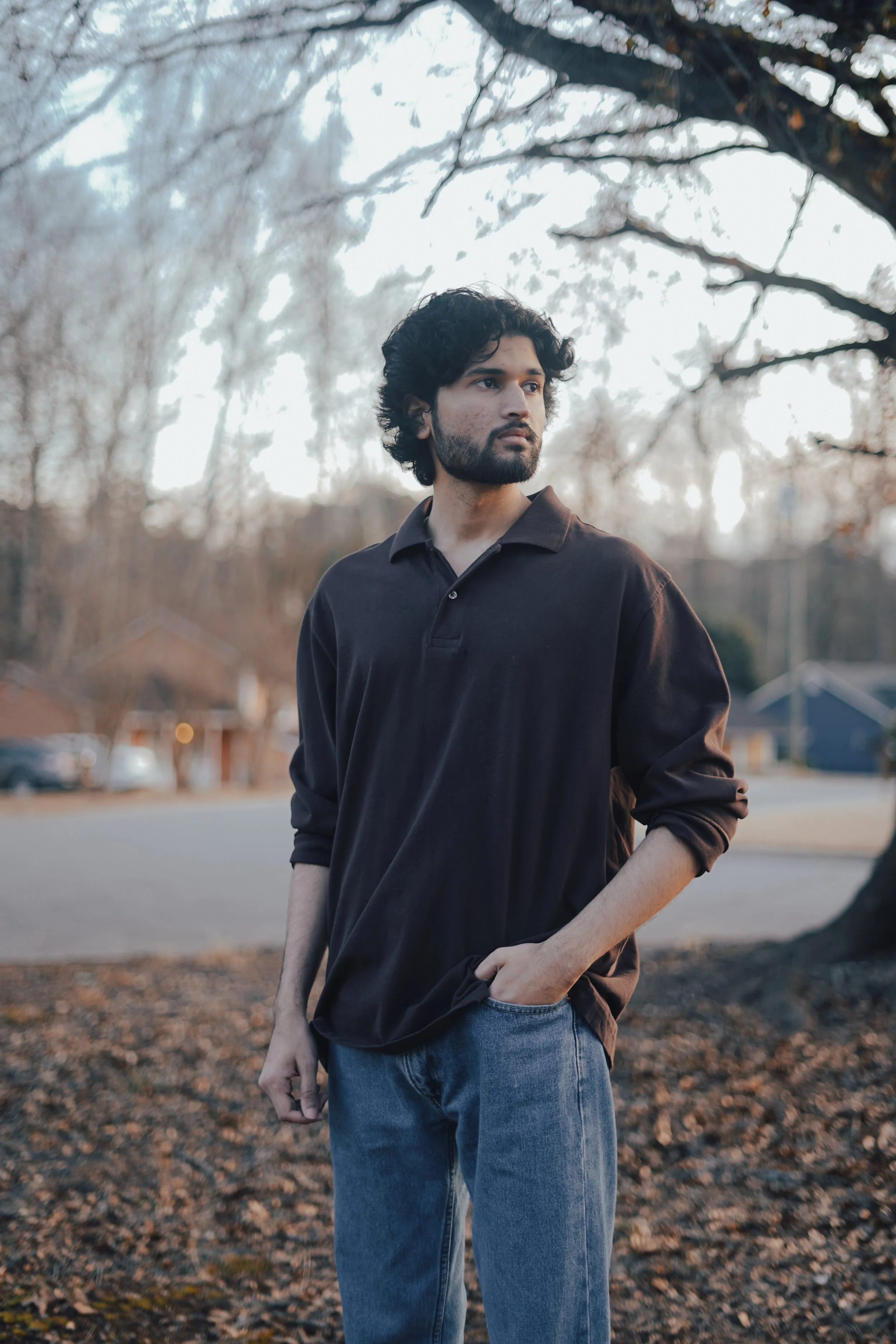 A young man with curly black hair and a beard, wearing a black long-sleeve shirt and blue jeans, standing outdoors in a park during autumn, with bare trees and a residential neighborhood in the background.