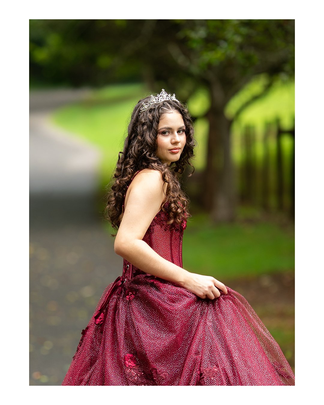 Young woman in a burgundy gown and tiara standing outdoors on a tree-lined path.
