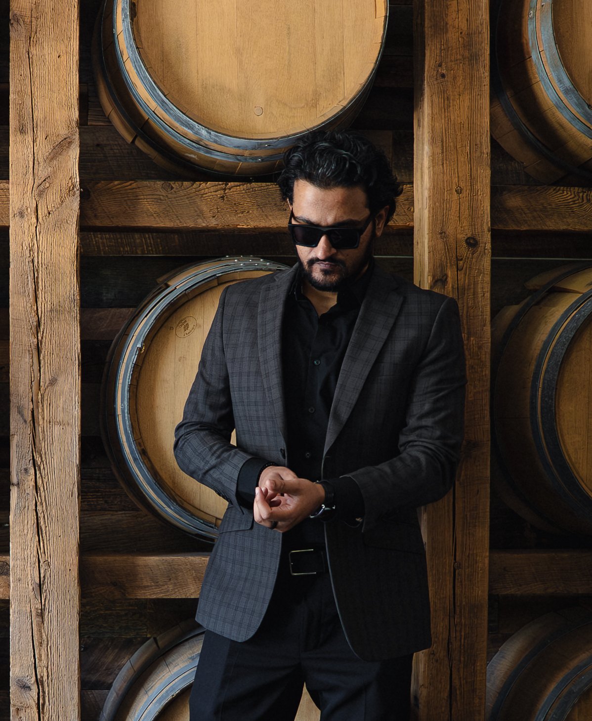 Man in a checkered gray suit and black shirt, wearing black sunglasses, standing in front of wine barrels made of wood.