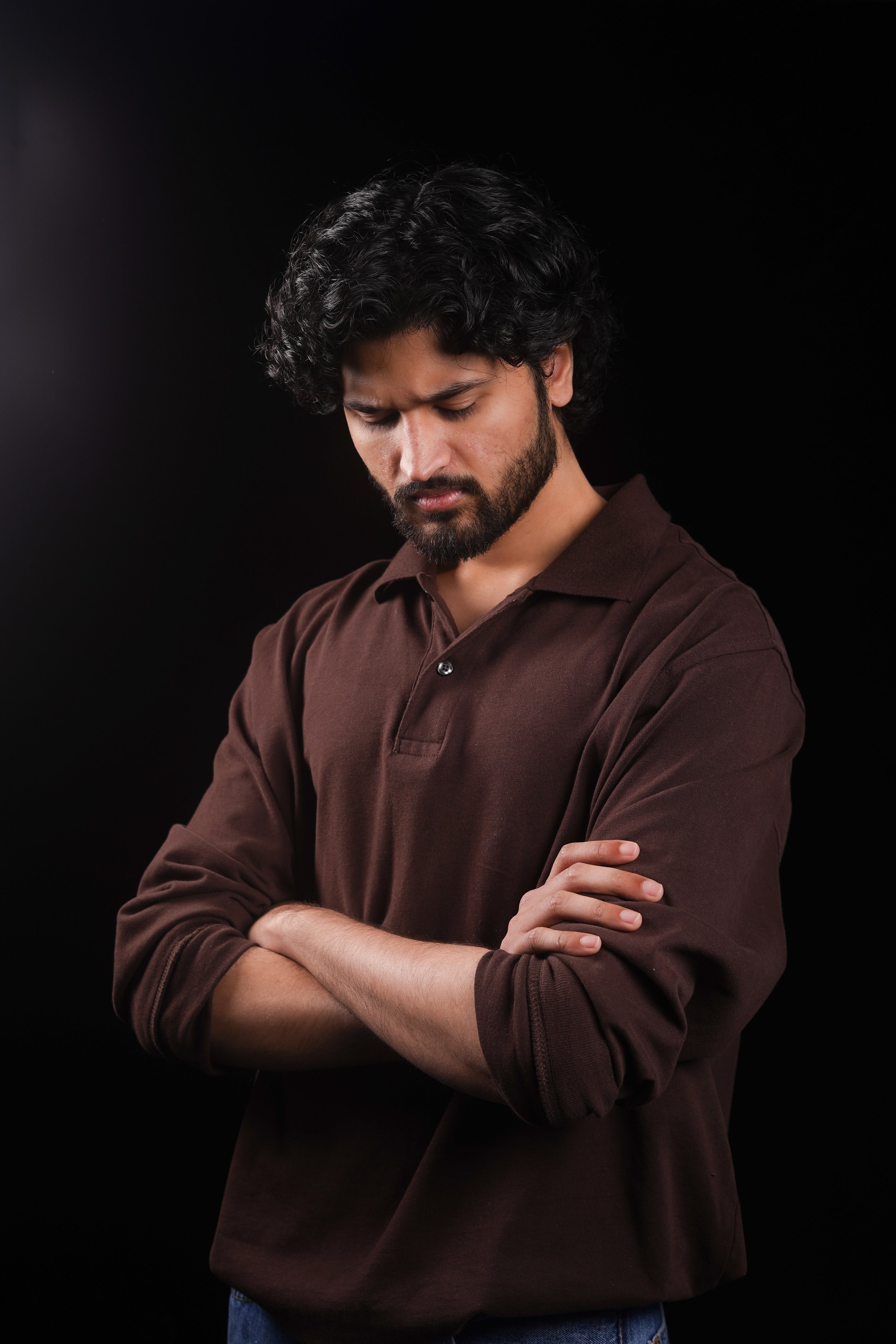A man with dark hair and a beard wearing a brown long-sleeve shirt with crossed arms, looking down against a black background.