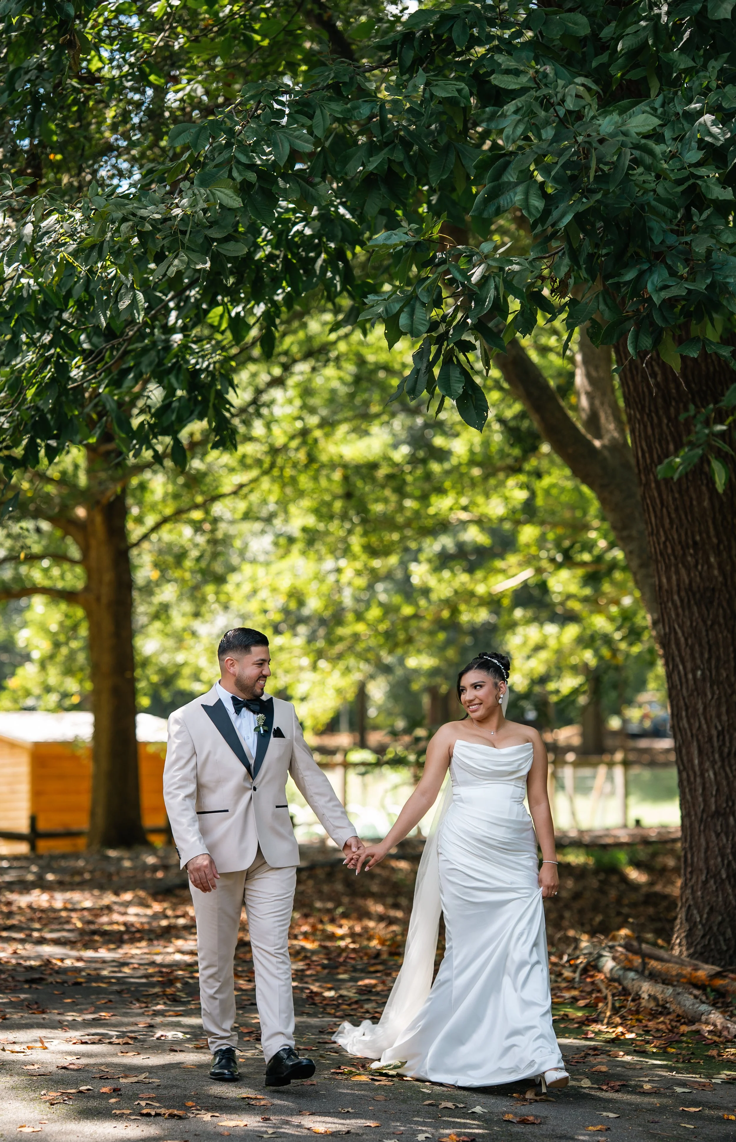 A newlywed couple holding hands and walking outdoors on a leaf-covered path surrounded by trees. The groom is wearing a white tuxedo and the bride is in a strapless white wedding dress. They are smiling and looking at each other.