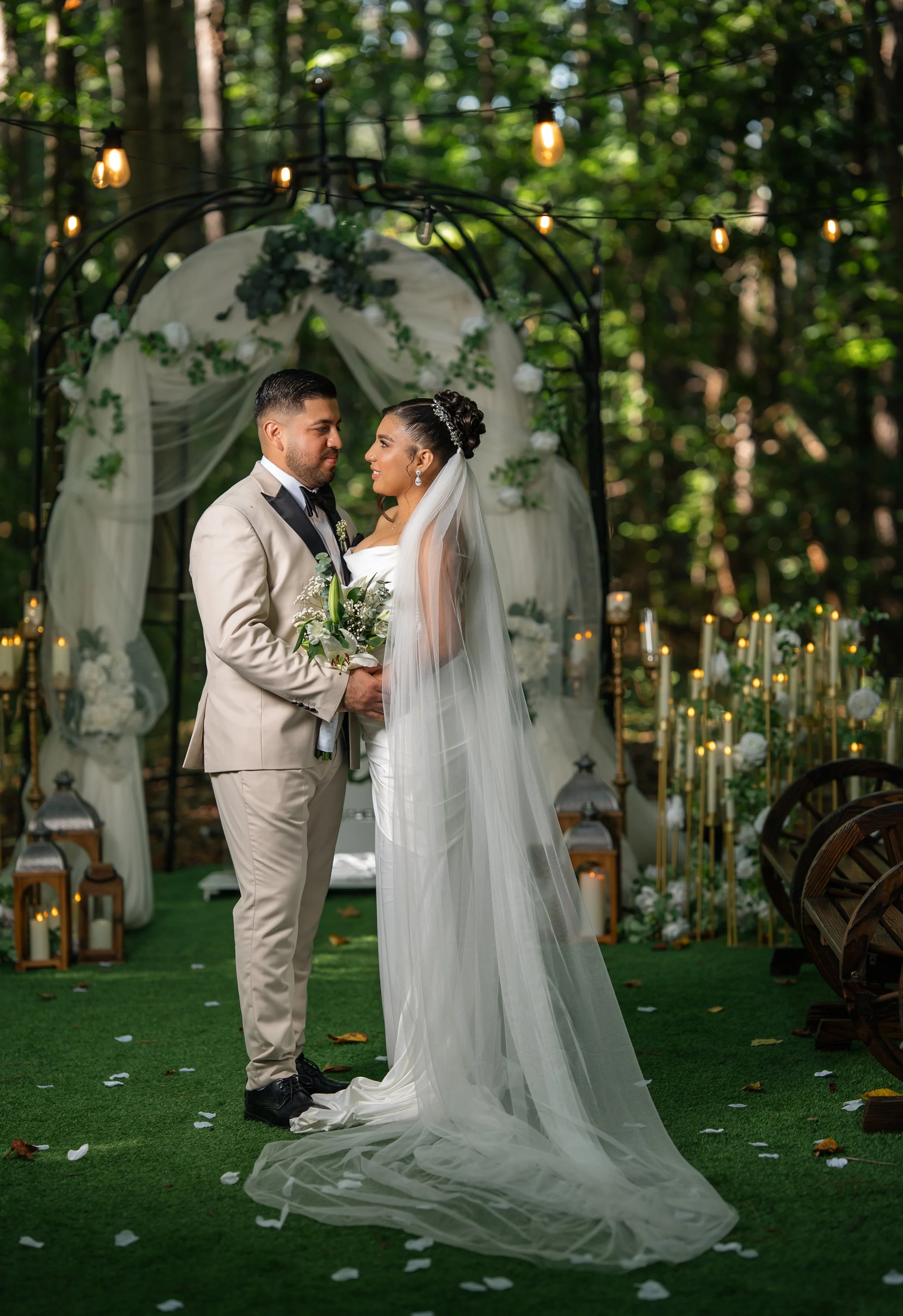 A bride and groom standing face to face during their outdoor wedding ceremony in a wooded area. The bride is wearing a white strapless wedding gown with a long veil, and the groom is wearing a beige tuxedo with a black bow tie. The setting is decorat
