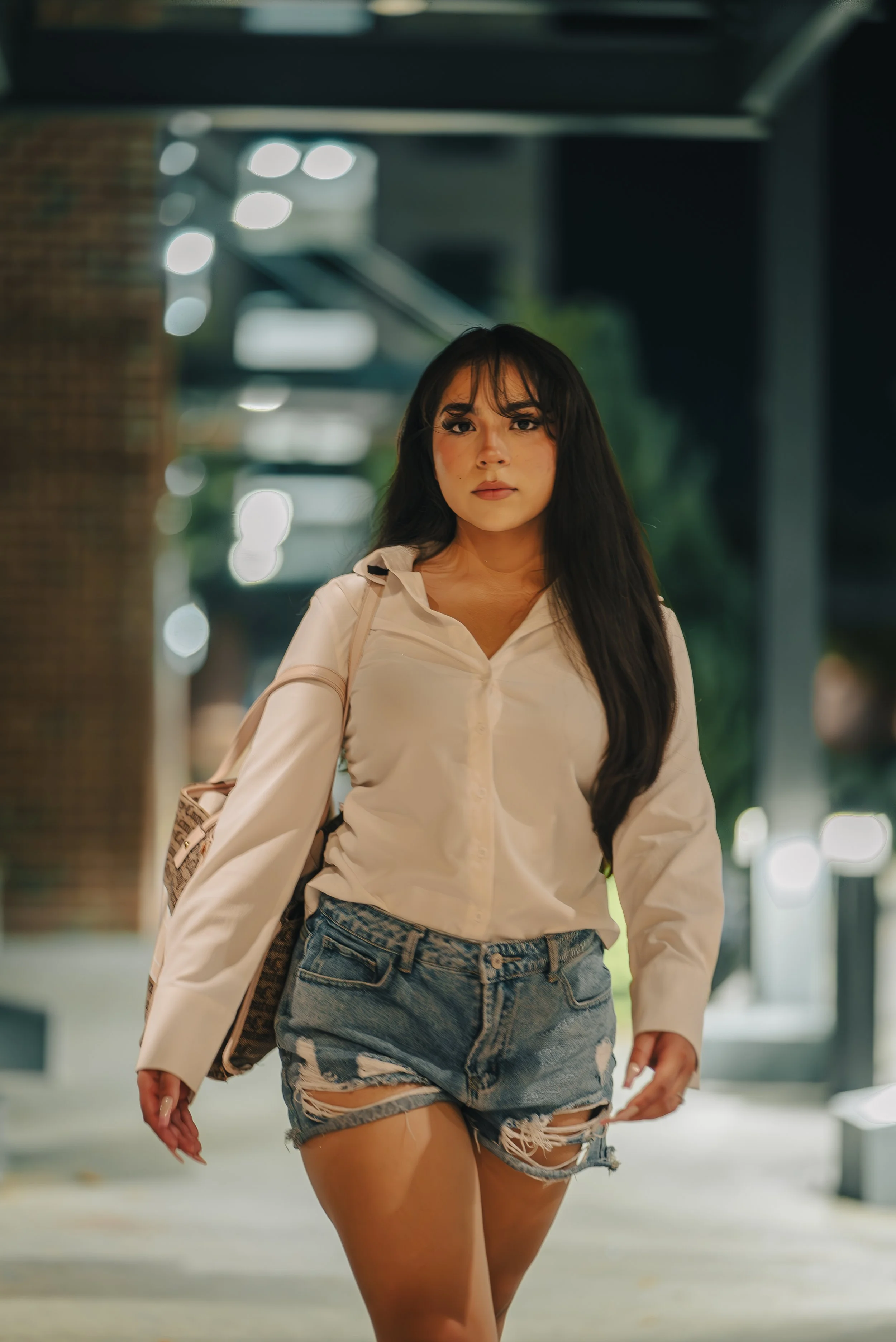 A woman with long dark hair, wearing a white button-up shirt and distressed denim shorts, walking outdoors at night.