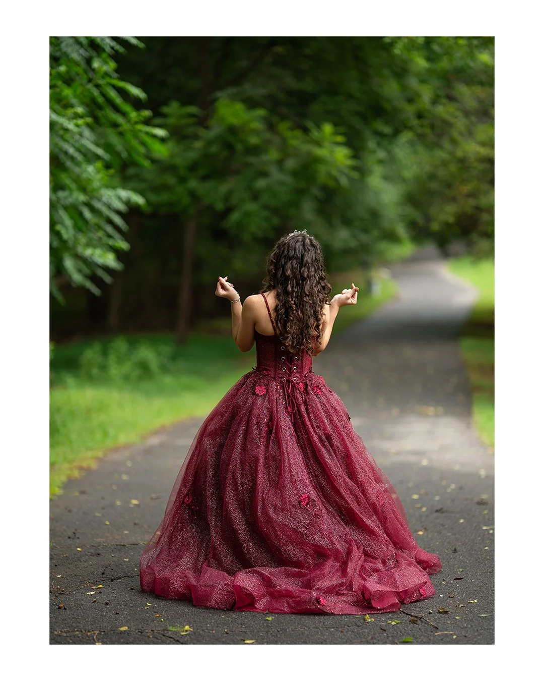 A woman in a burgundy gown standing on a paved road in a green wooded area, facing away, with her hands raised in a meditation pose.