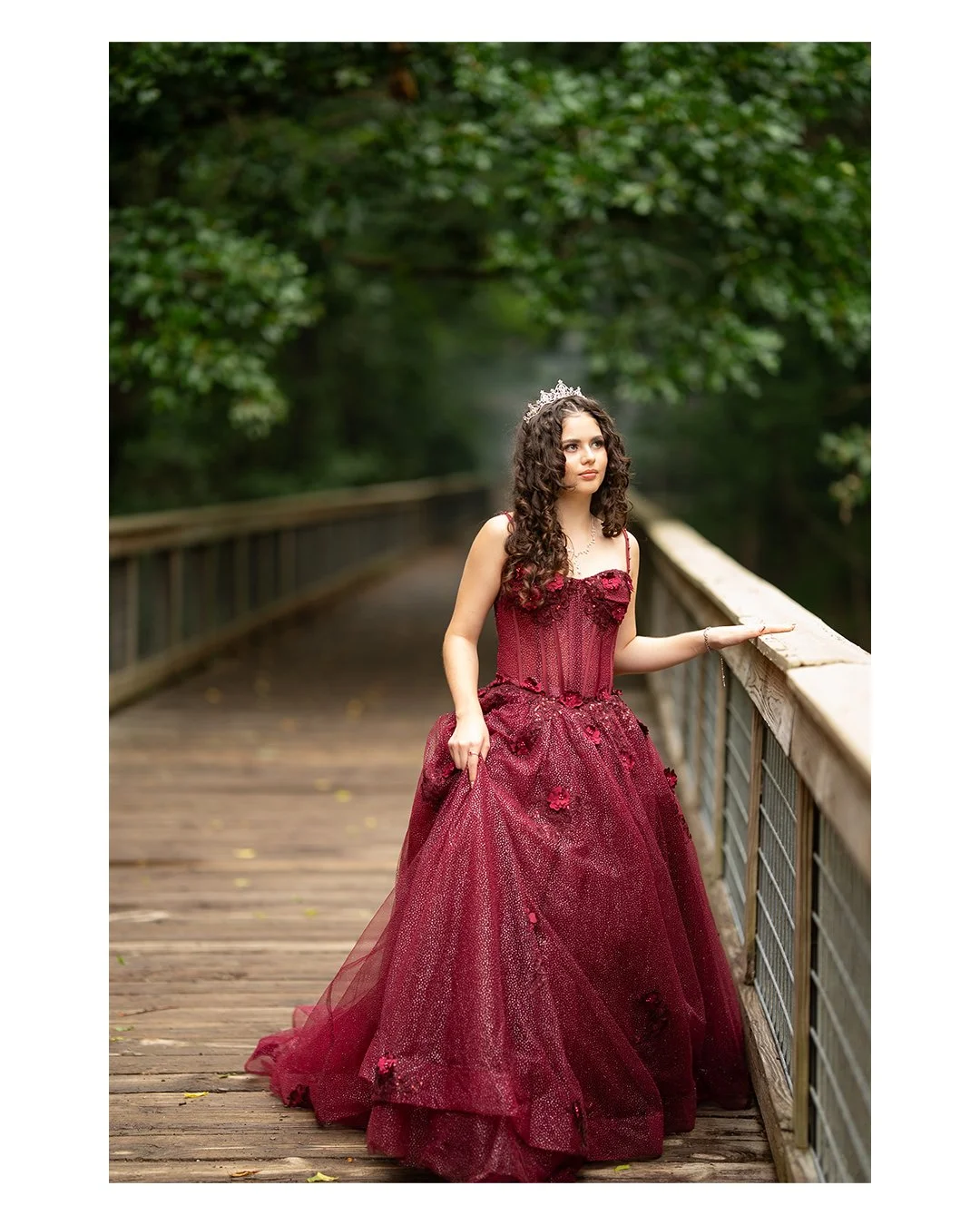 Young woman with curly hair wearing a crimson ball gown and a tiara, standing on a wooden bridge surrounded by lush green foliage.