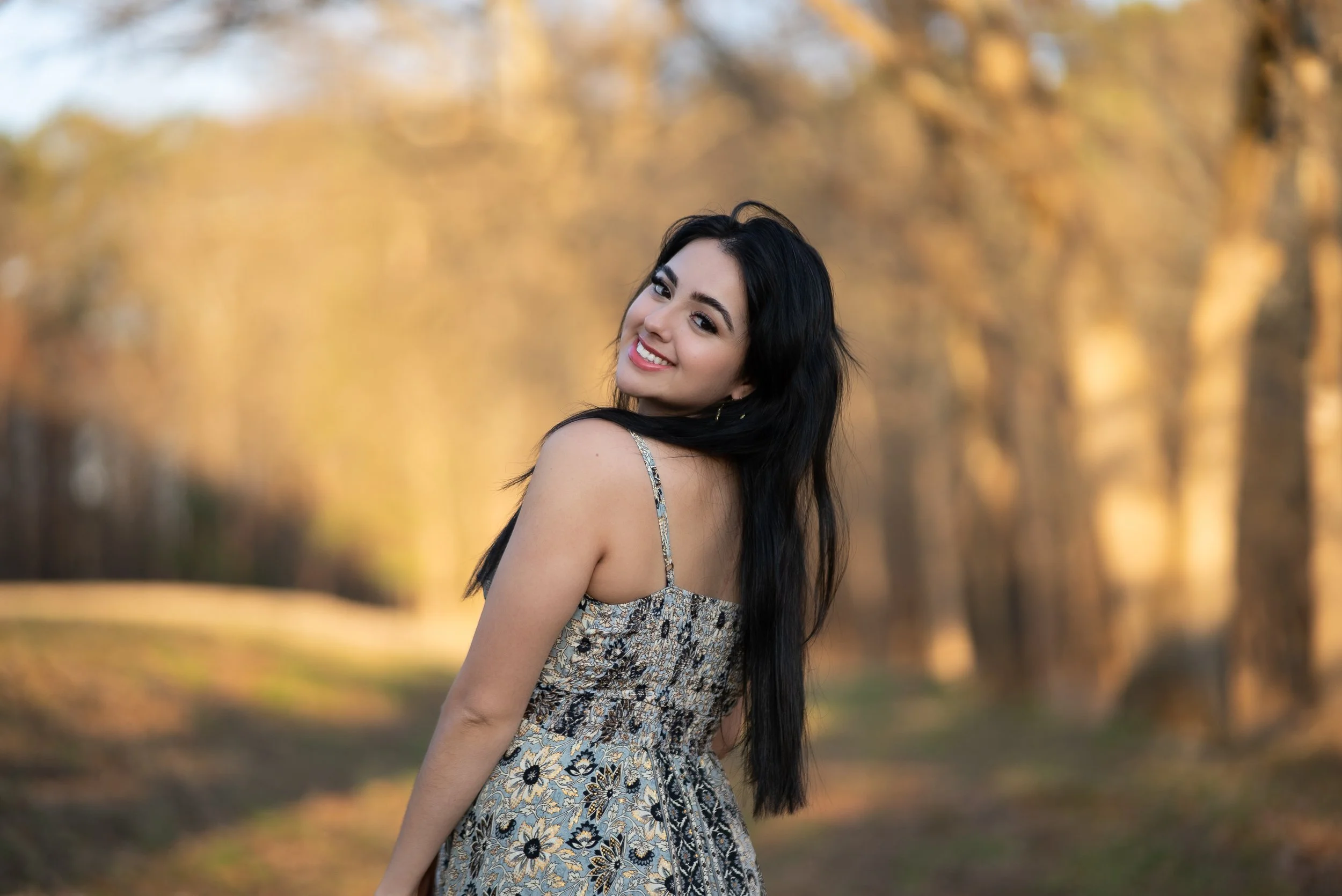 Smiling young woman with long dark hair in a patterned dress outdoors, with autumn trees in the background.