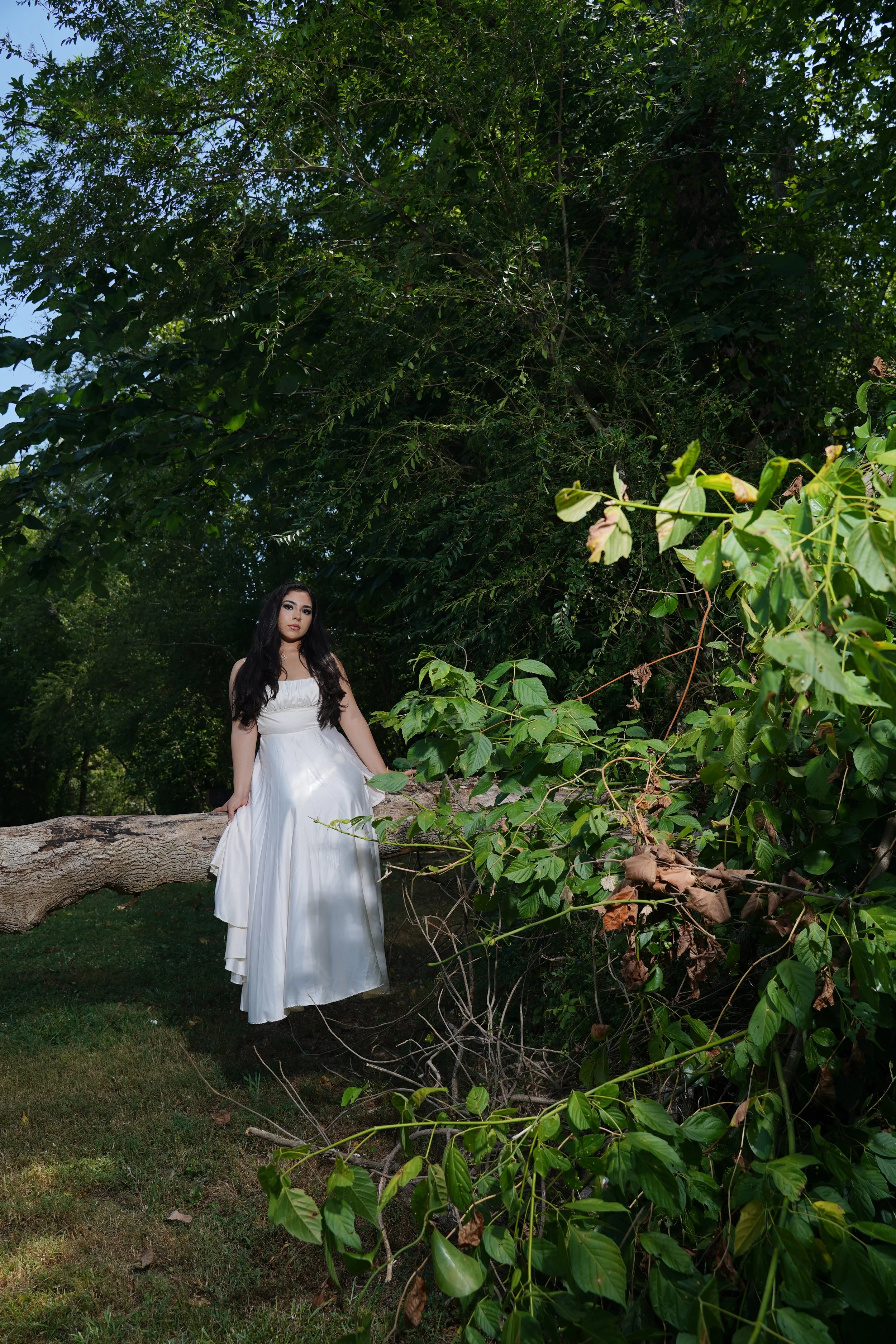 A woman in a long white dress sitting on a fallen tree trunk in a lush green outdoor setting with dense foliage.