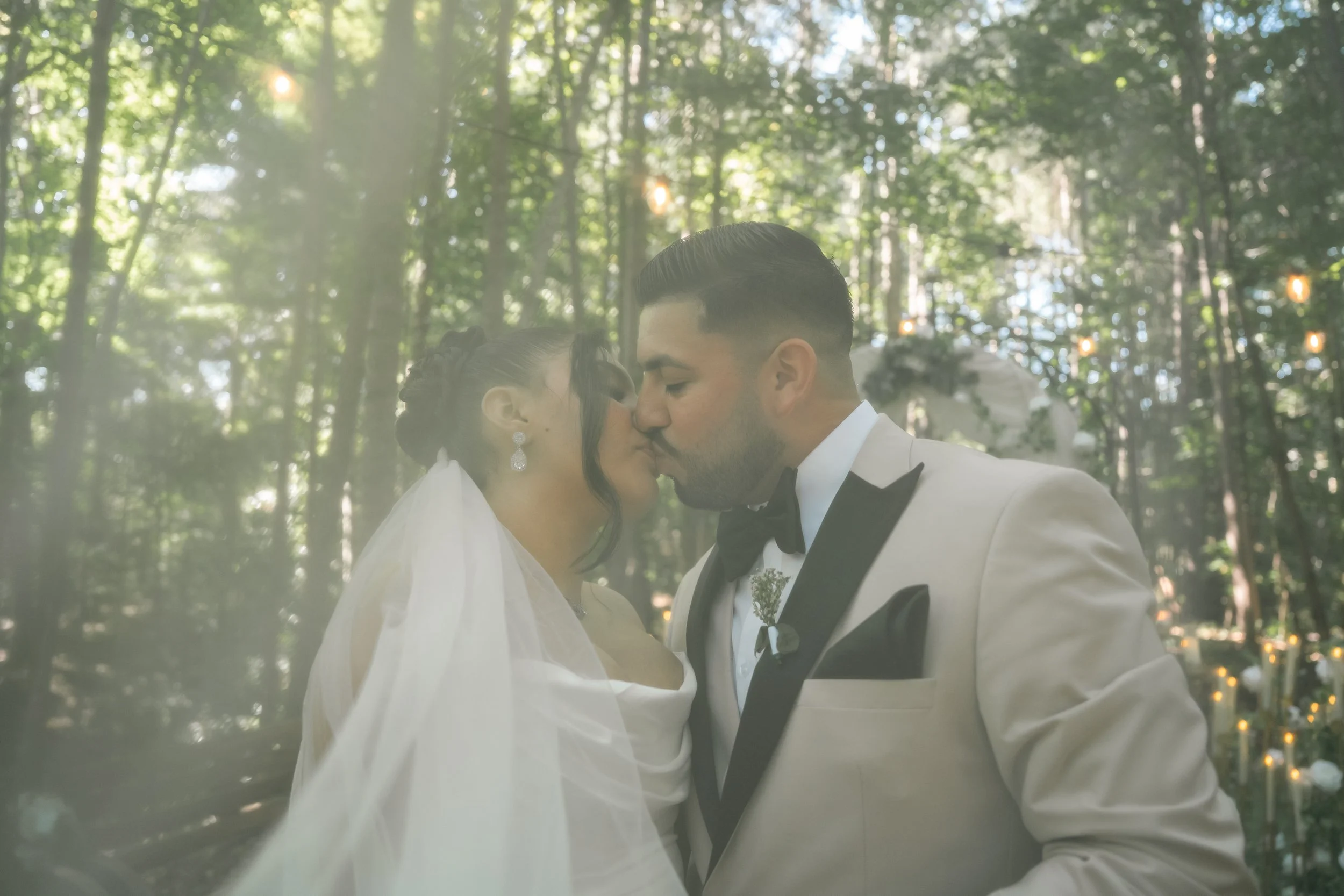 A couple dressed in wedding attire, with the bride wearing a veil and wedding earrings, and the groom wearing a white tuxedo jacket with black lapels and a bow tie, sharing a close moment in a forest setting with hanging lights in the background.
