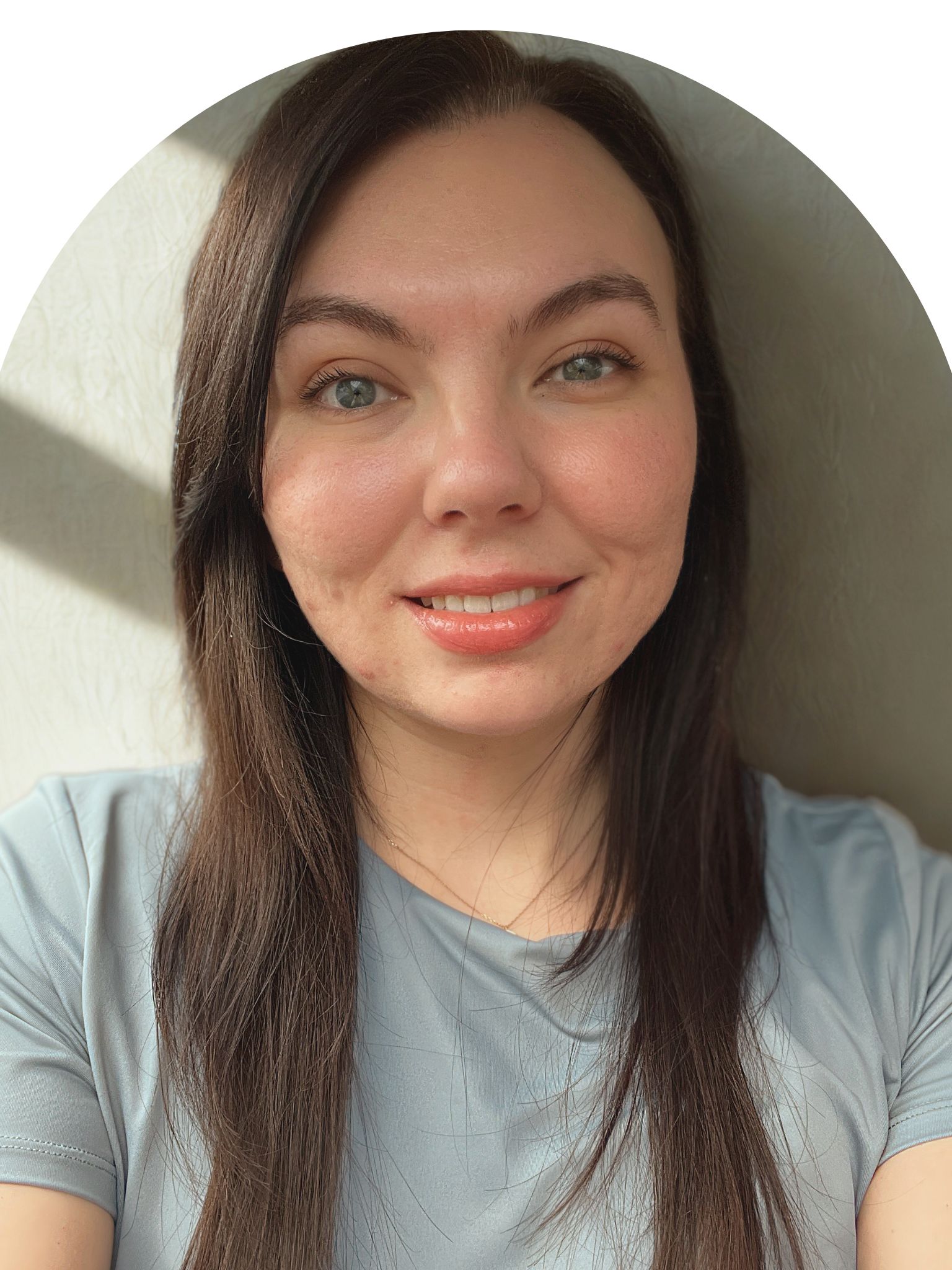 A woman with long brown hair and blue eyes smiling in front of a light-colored textured wall, partially illuminated by sunlight.