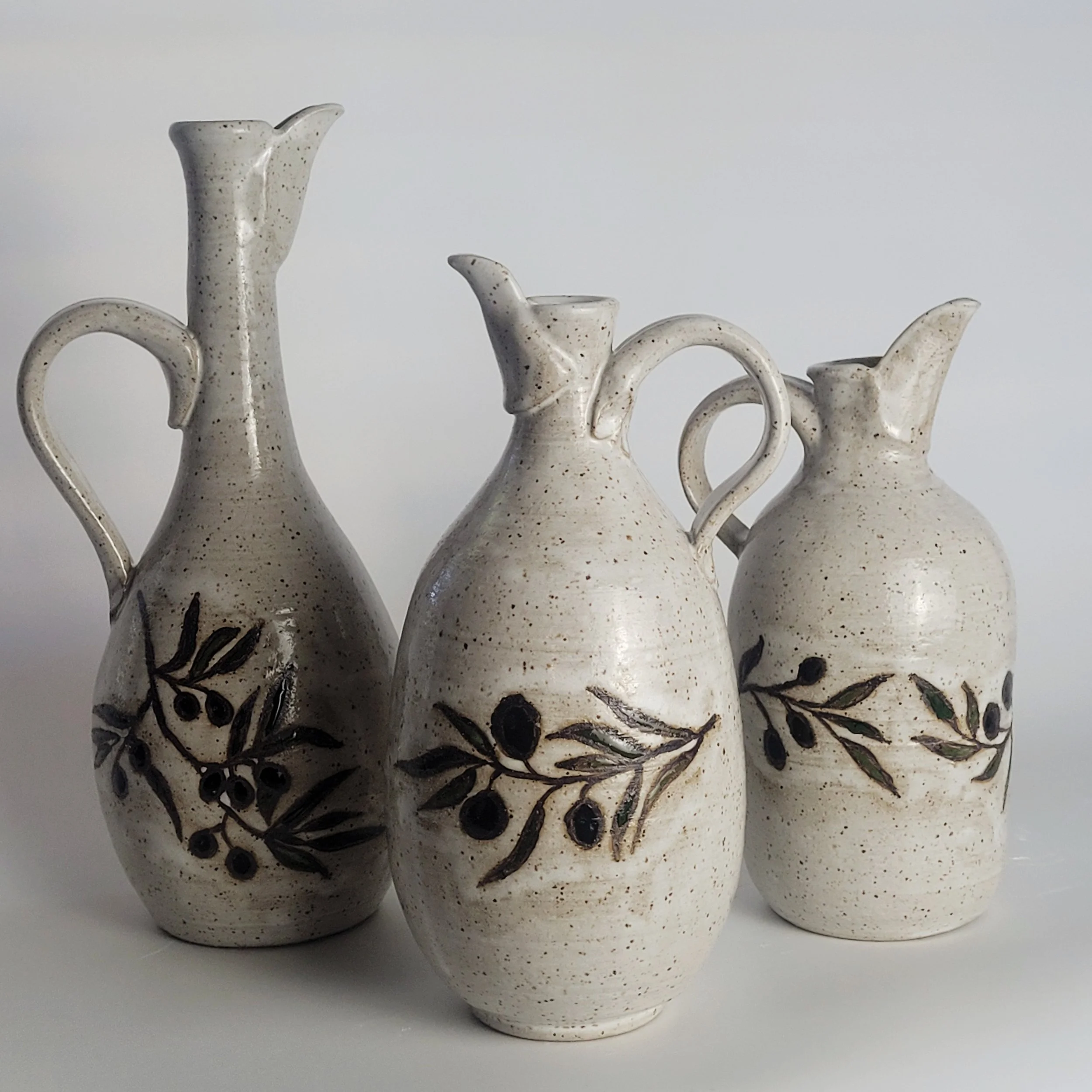 Three ceramic vases with olive branch and olive designs painted on them, all with a speckled cream glaze, placed against a plain background.