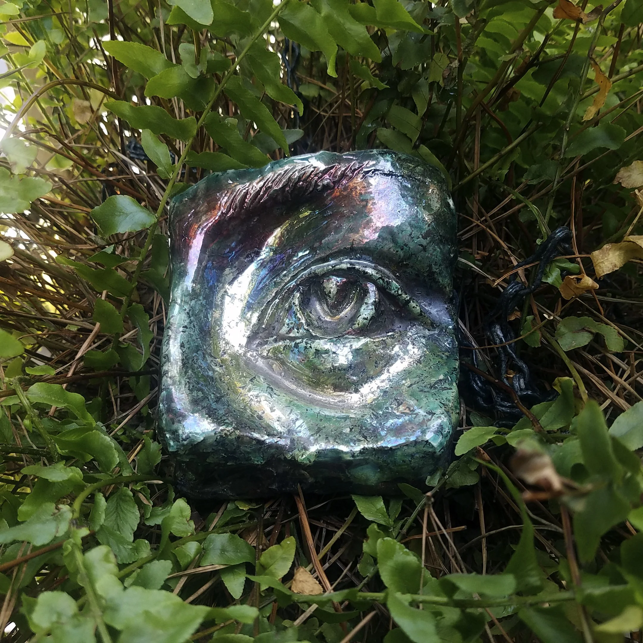 A metallic or ceramic sculpture of an eye, designed with a reflective, iridescent surface, surrounded by green leafy plants and brown vines.