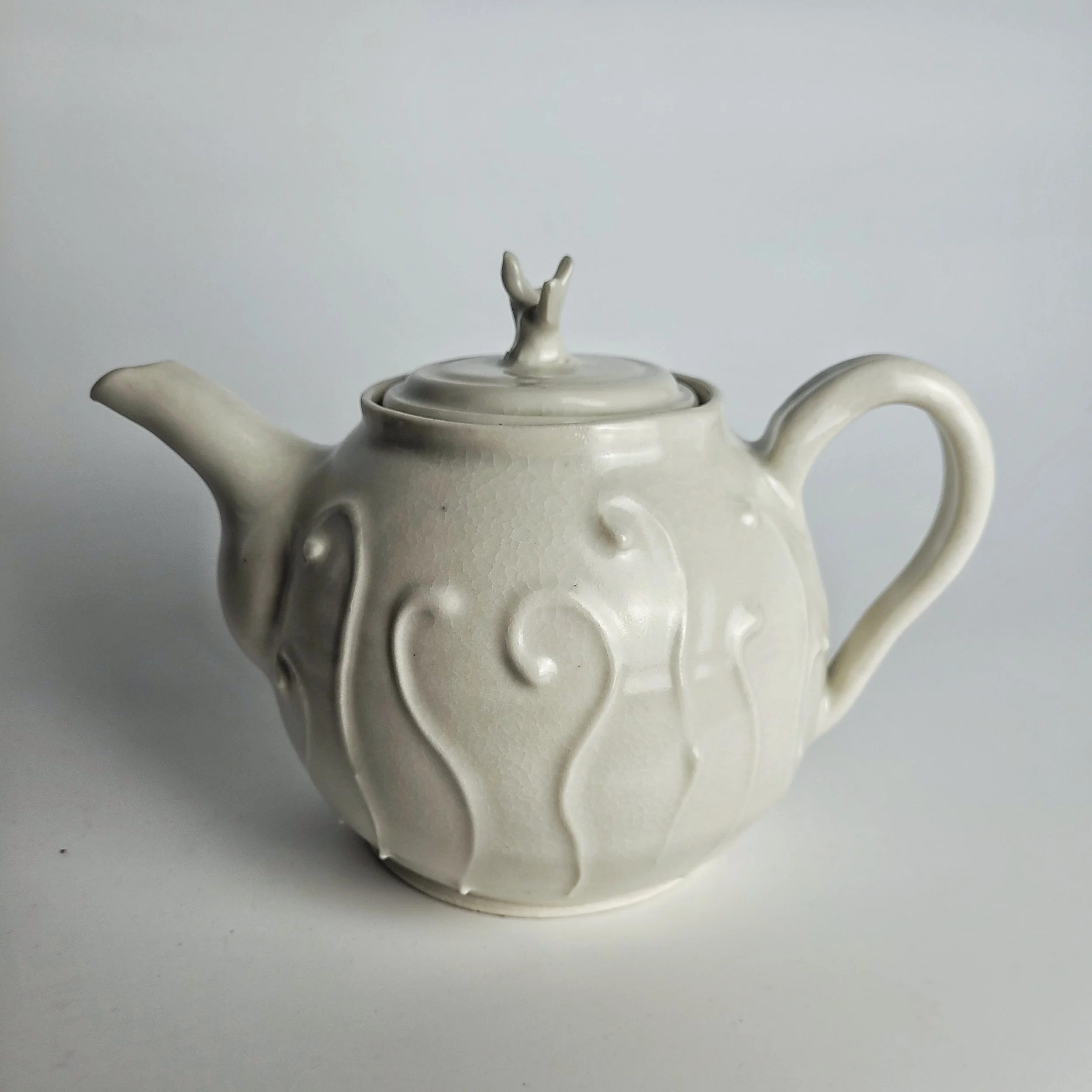 White ceramic teapot with embossed swirl and line patterns, a curved handle, a spout, and a lid with a small decorative handle on top, set against a plain light background.