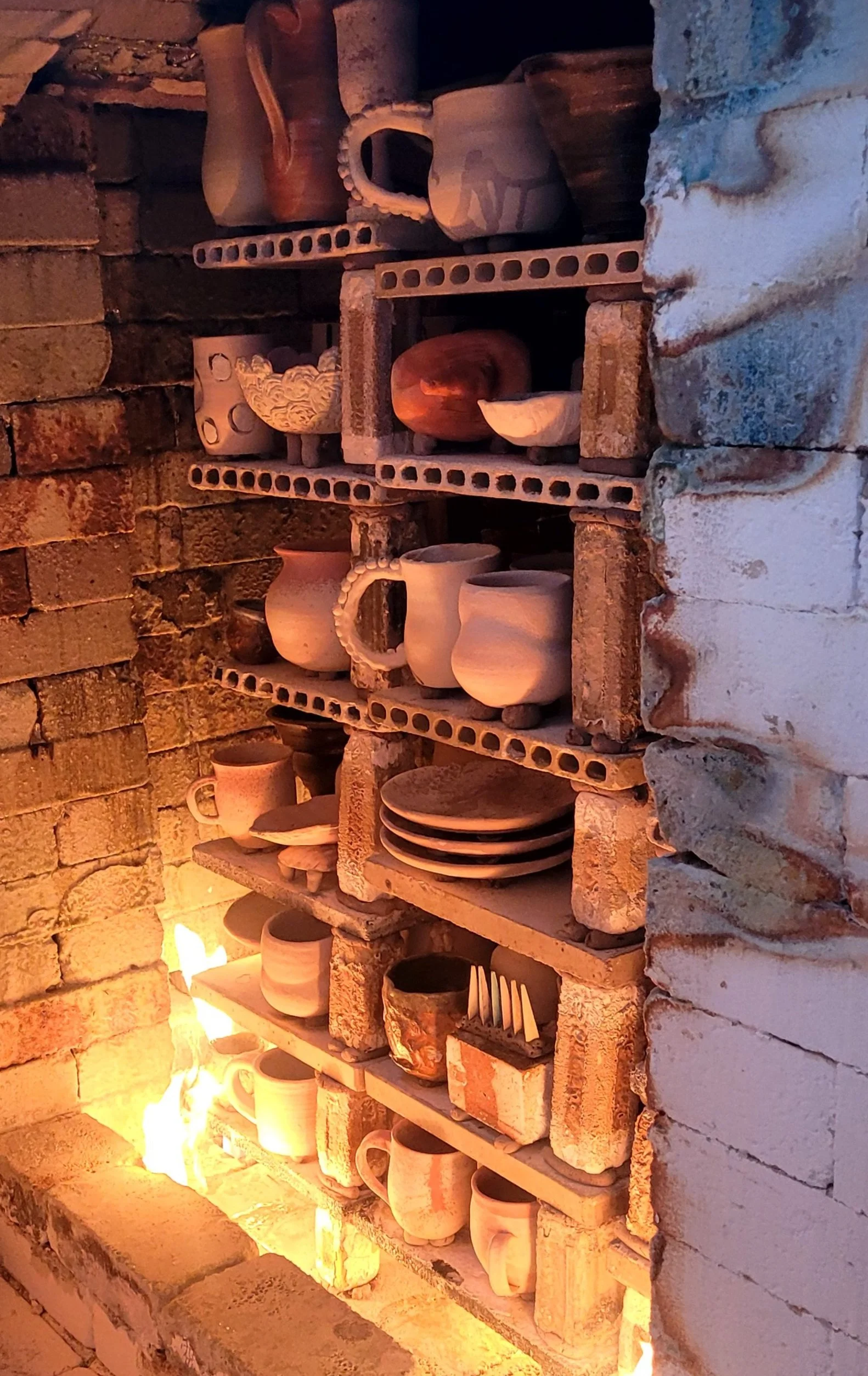 Shelves filled with various pottery and ceramic cups, bowls, and plates, set against a brick wall with a fire glowing at the bottom.