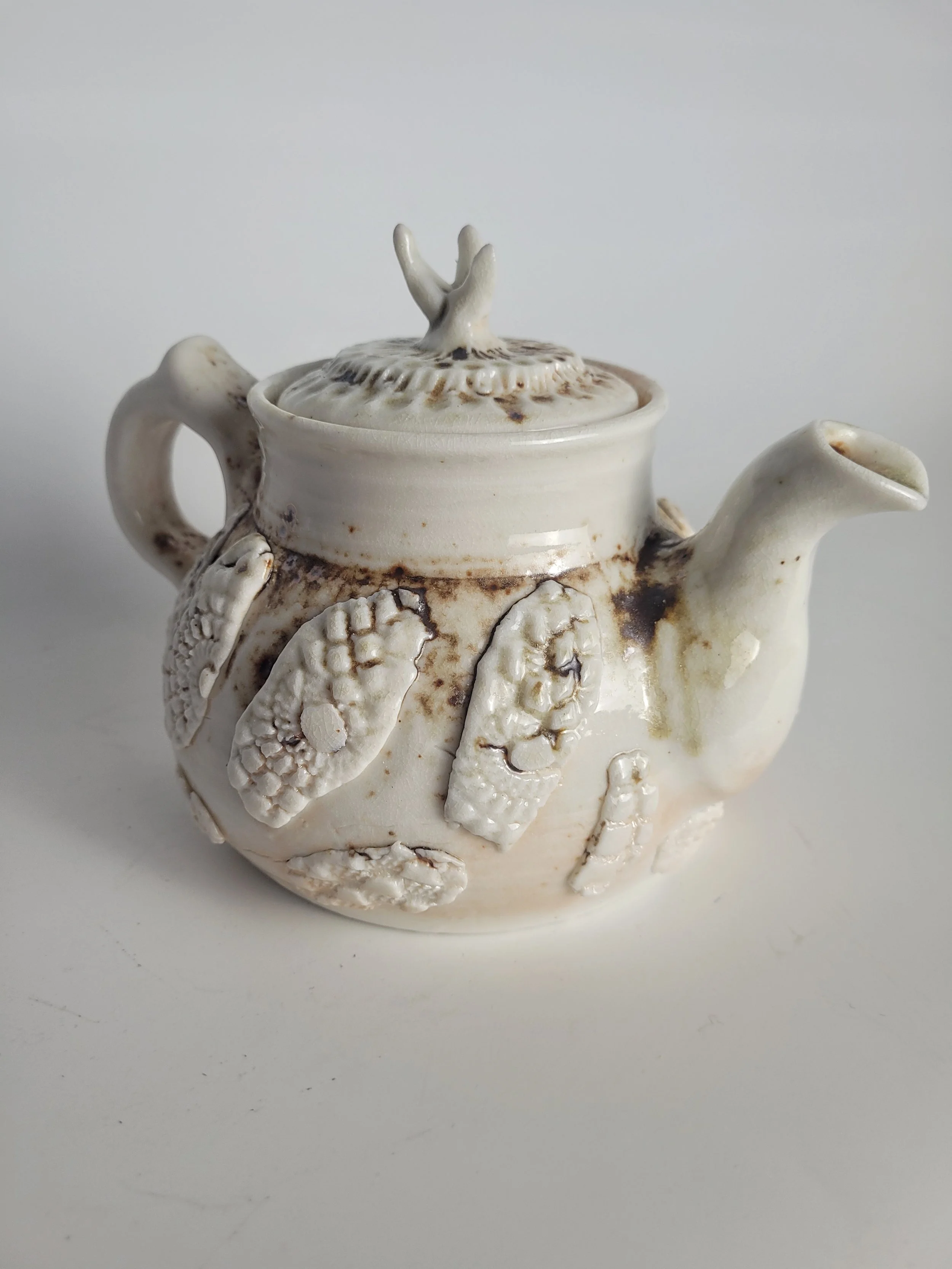 A ceramic teapot with textured, raised designs of fish on its body and a lid featuring a small, decorative hand-shaped handle.