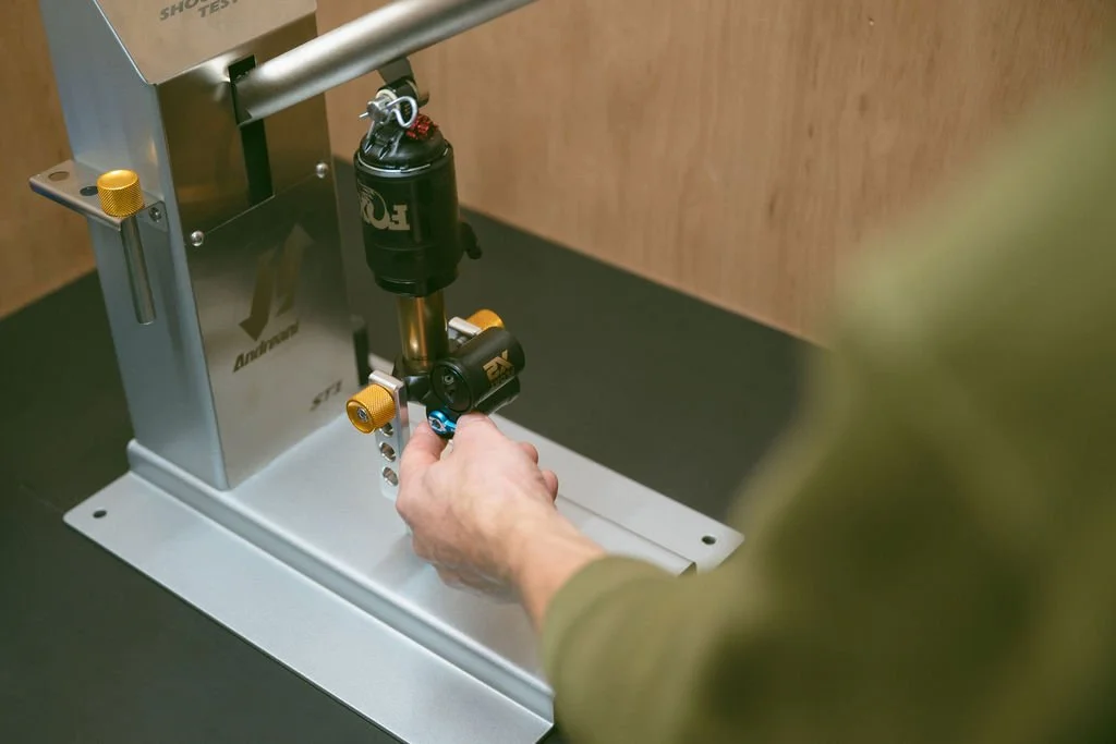 Close-up of a person's hand adjusting suspension components on a motorcycle test stand, with a black shock absorber and yellow adjustment knobs.