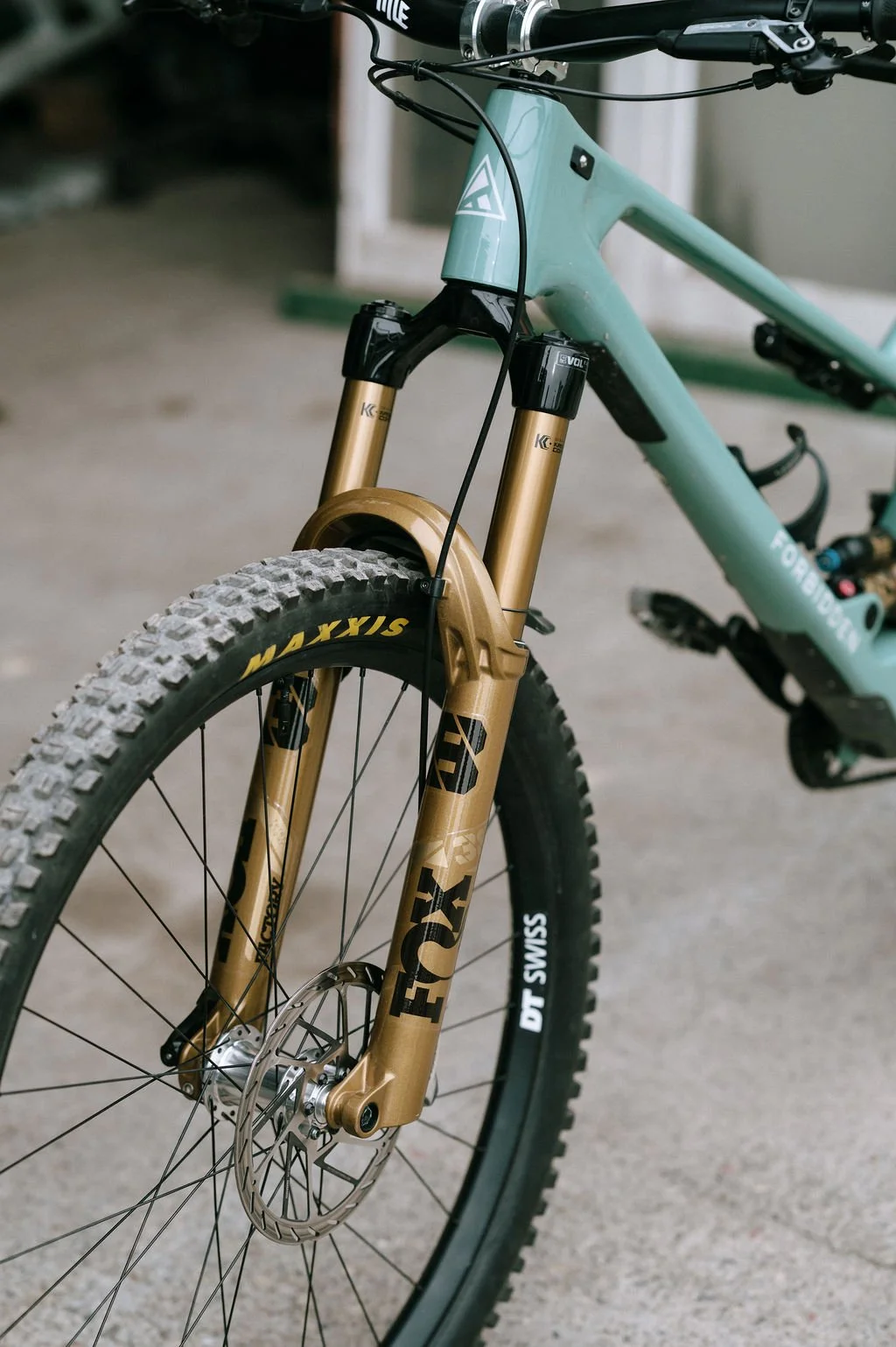 Close-up of a teal mountain bike with gold suspension fork, disc brakes, and knobby tires, parked on a concrete surface.