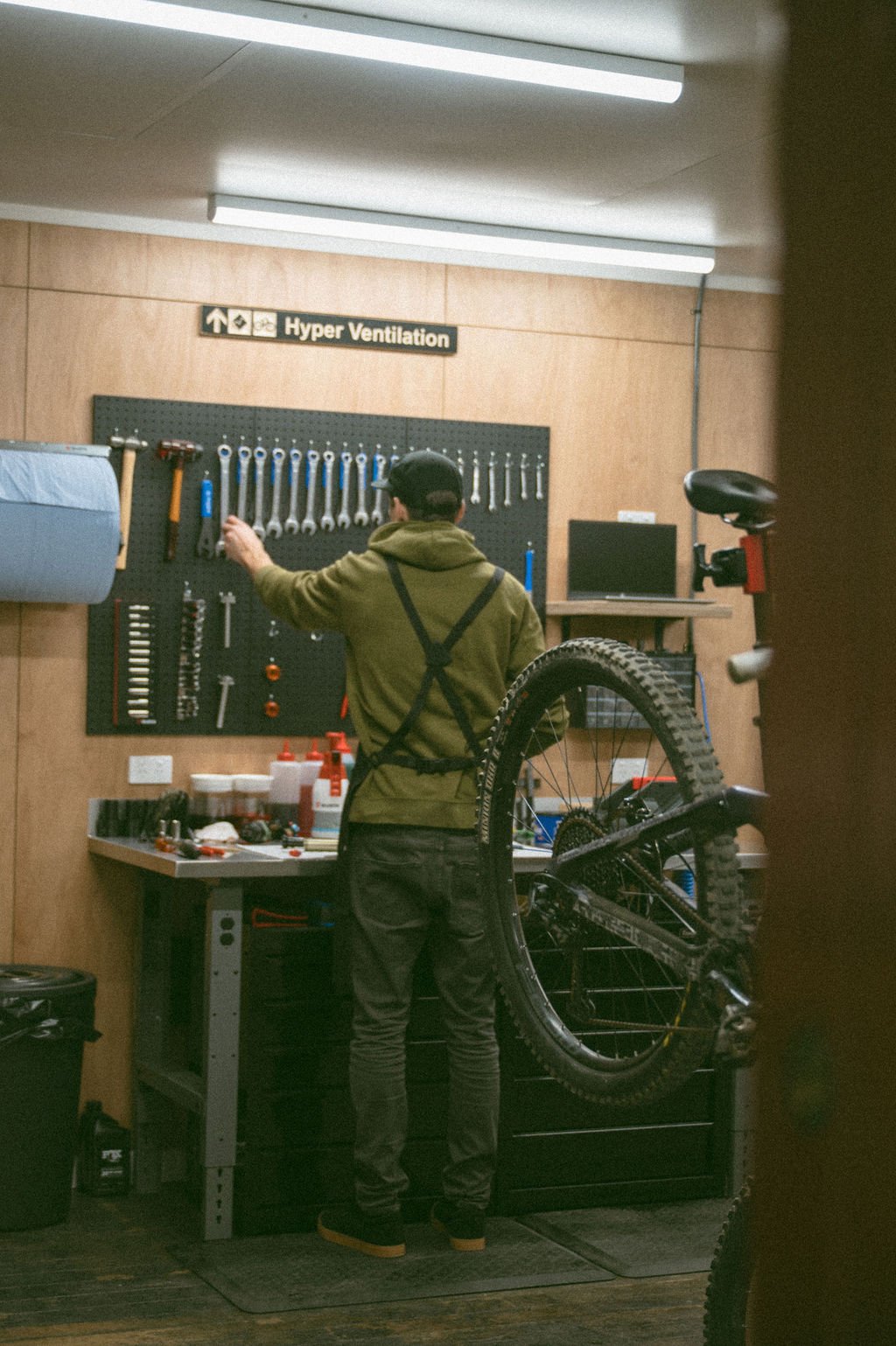 A person working on bike maintenance in a workshop with a wooden wall, pegboard tools, and a 'Hyper Ventilation' sign above.