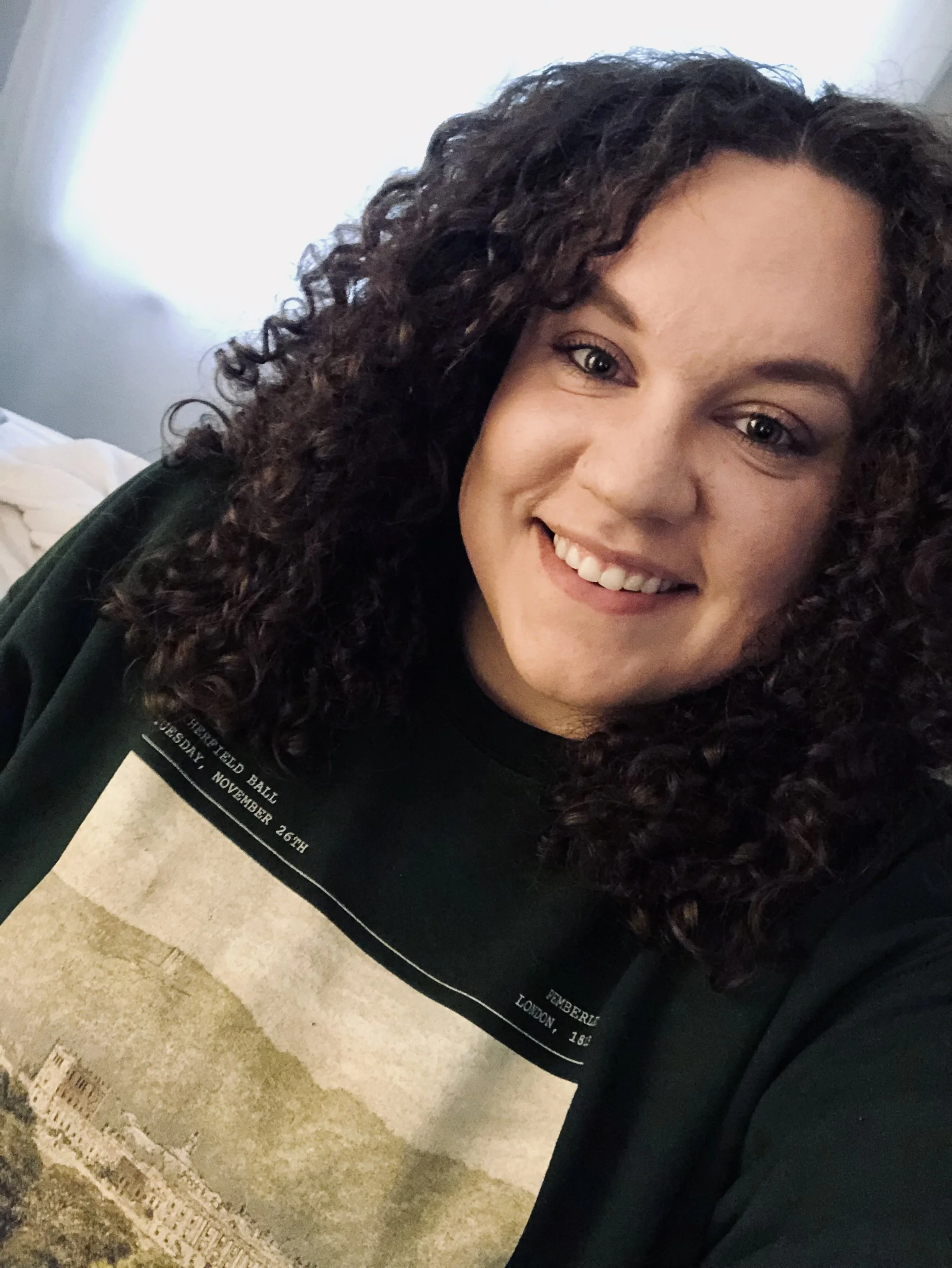 A woman with dark, curly hair smiling in a close-up selfie, wearing a black T-shirt with a sepia-toned image of a cityscape and some text.