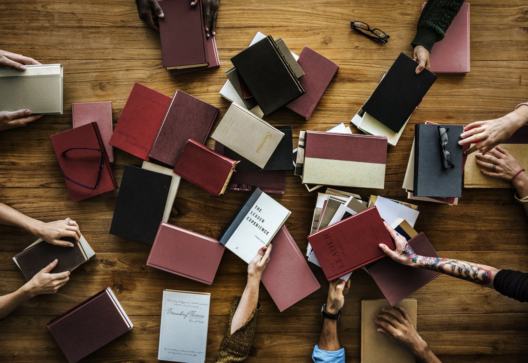 Pile of books on a large wooden take viewed from the top with many people's hands around the table reaching for the books